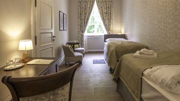 A hotel room with a desk, two beds, a chandelier, a window with curtains, and a patterned wall.