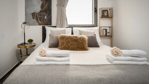 A neatly made bed with white and brown pillows, a fluffy brown accent, and neatly folded towels. There is a small side table with a lamp and a picture frame. A large abstract painting is on the wall above the bed.