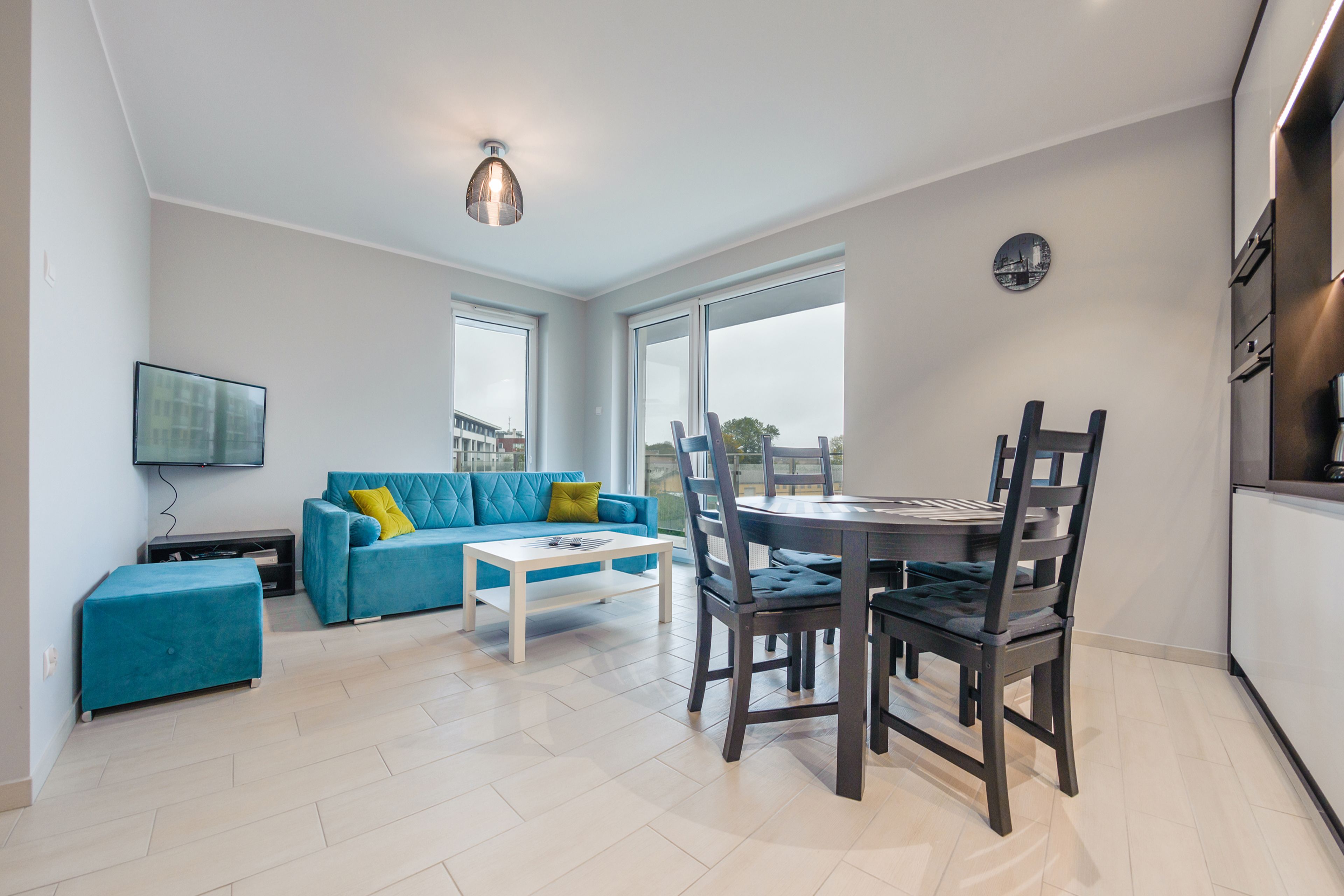 A modern living room with a blue sofa, a white coffee table, and a dining table with chairs. There is a flat-screen TV mounted on the wall and a cityscape view through the windows.