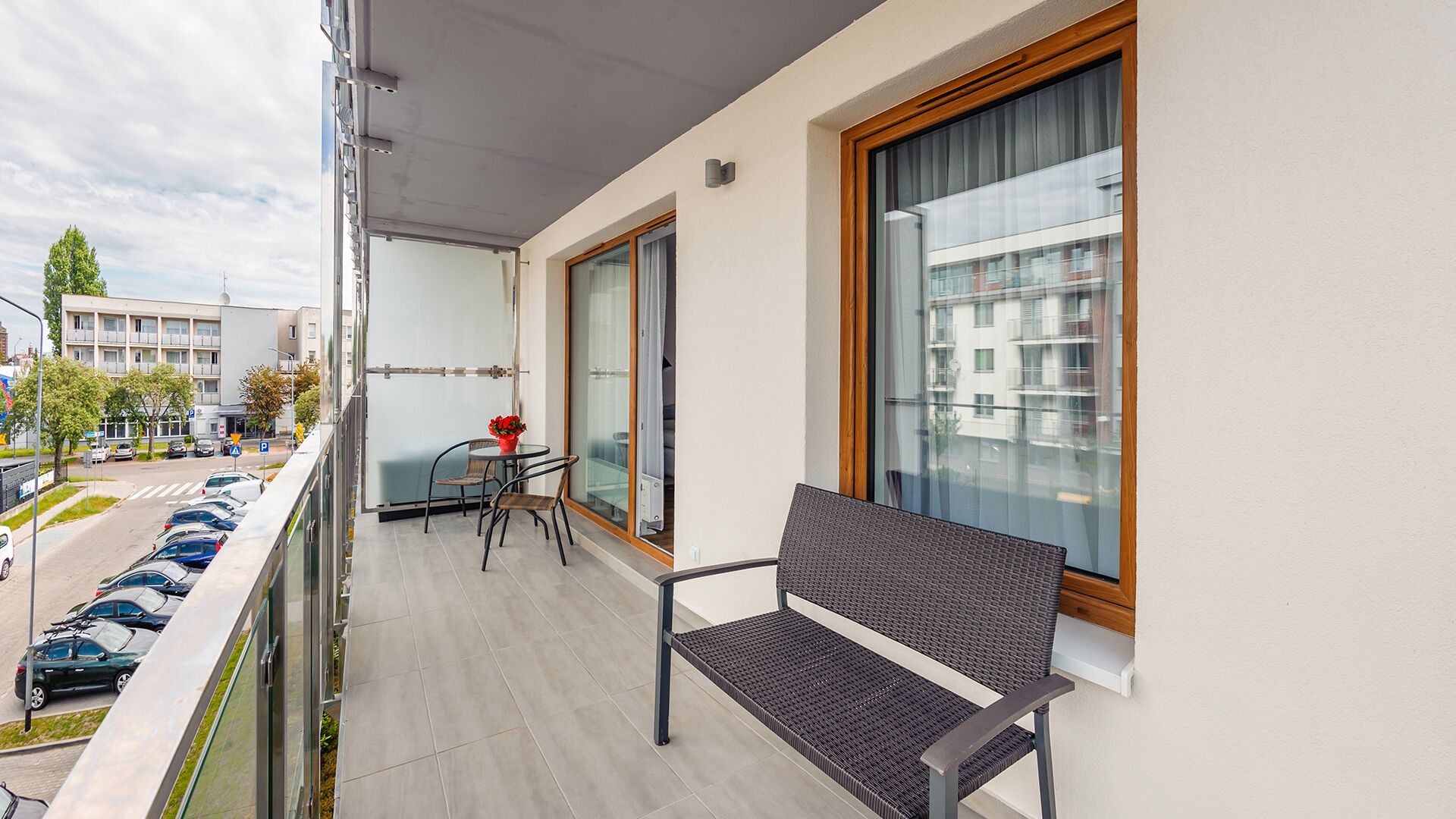 A balcony with a wicker chair, a small table with a vase, and a view of a street and buildings.