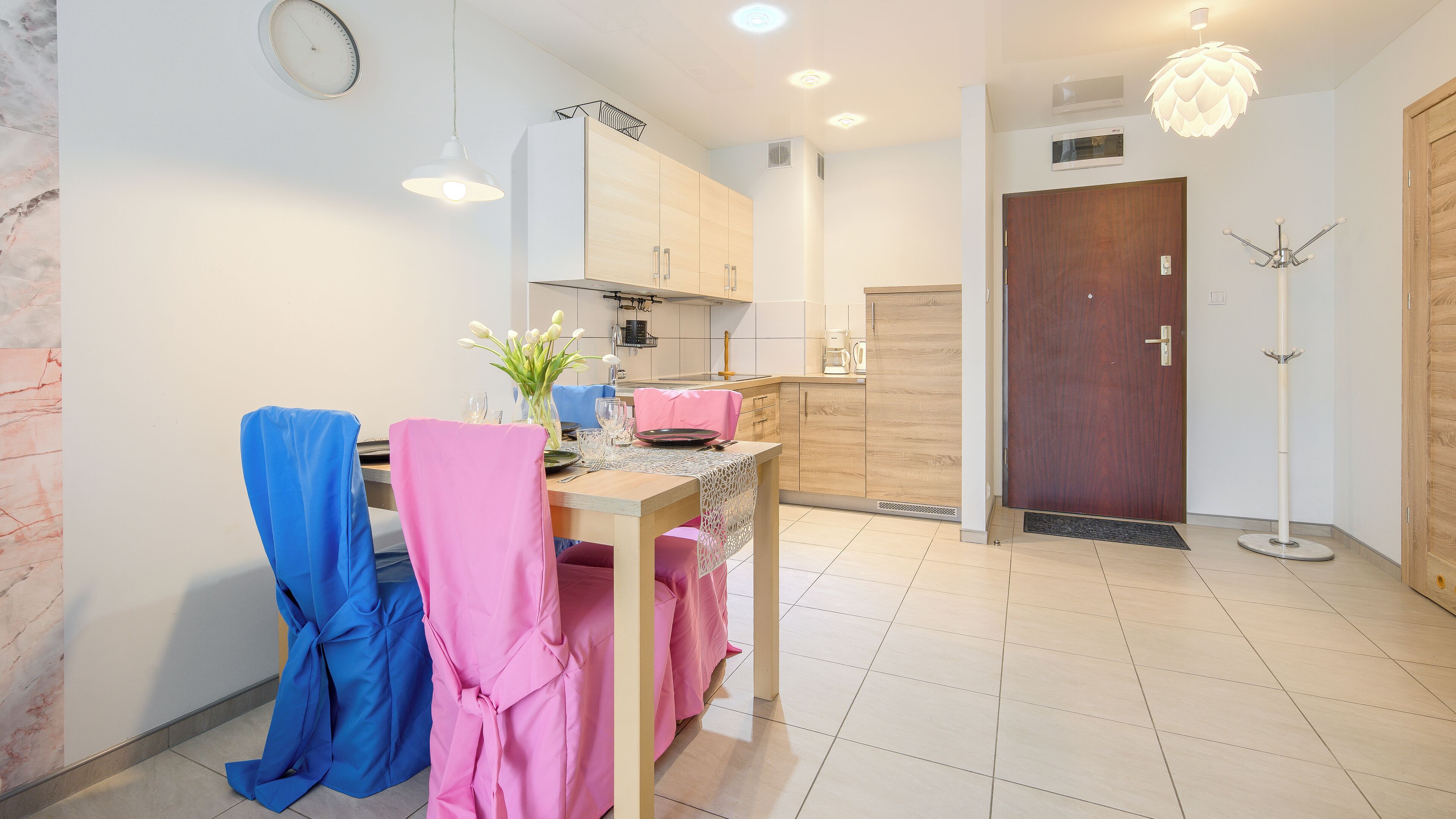 A modern kitchen with wooden cabinets, a dining table with pink and blue chairs, a vase with flowers, and a wooden door.