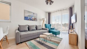 Un salon moderne avec un canapé gris, un fauteuil bleu, une table d’appoint en bois et une grande fenêtre avec des rideaux.