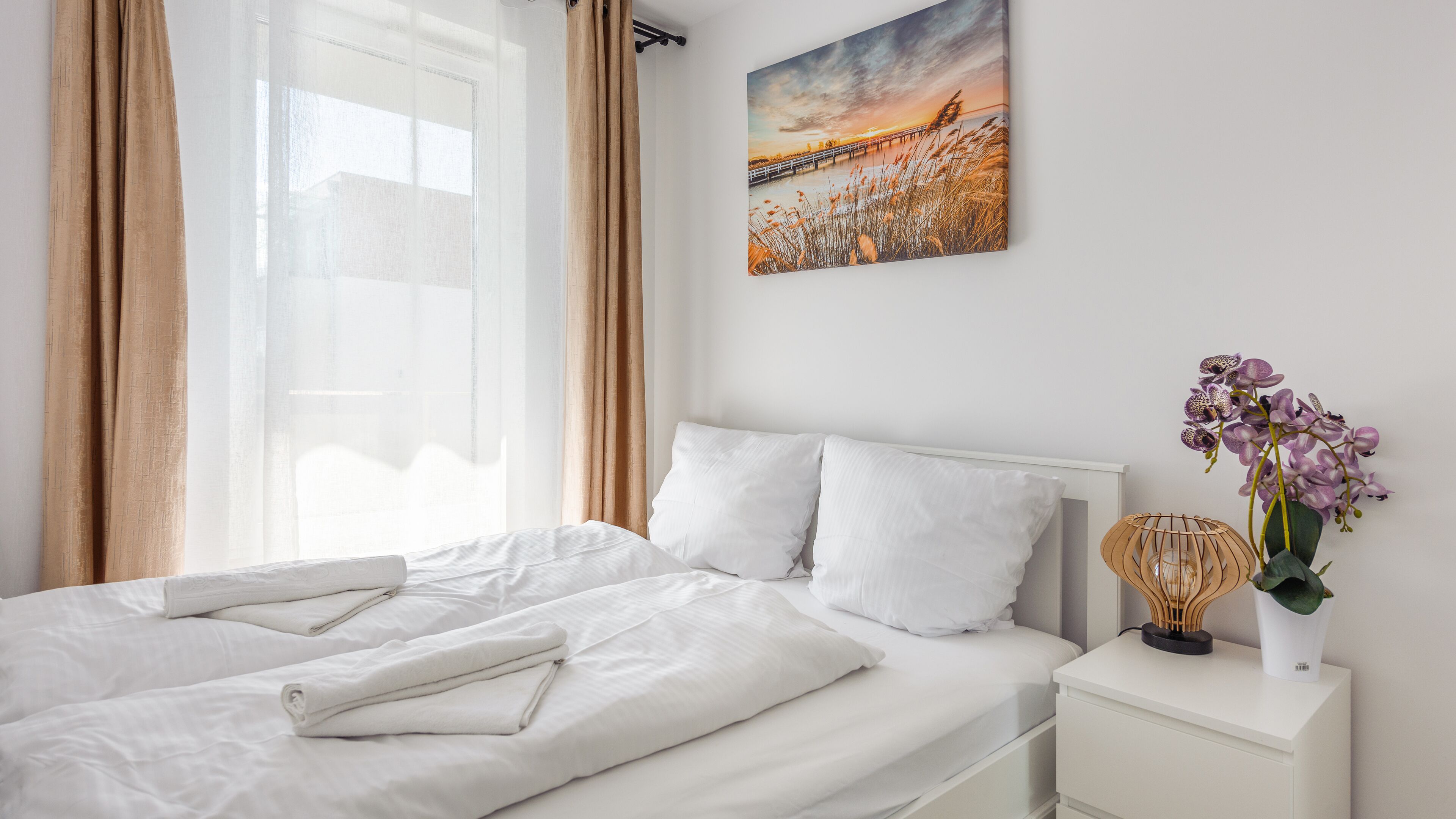 A neatly made bed with white linens and pillows, a painting of a sunset over a bridge on the wall, and a potted orchid on a bedside table.