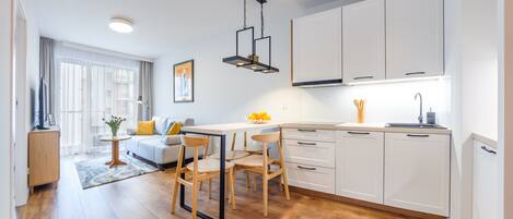 A modern kitchen with white cabinets, a wooden dining table, and chairs. There is a window with sheer curtains, a small side table with a vase, and a hanging light fixture.