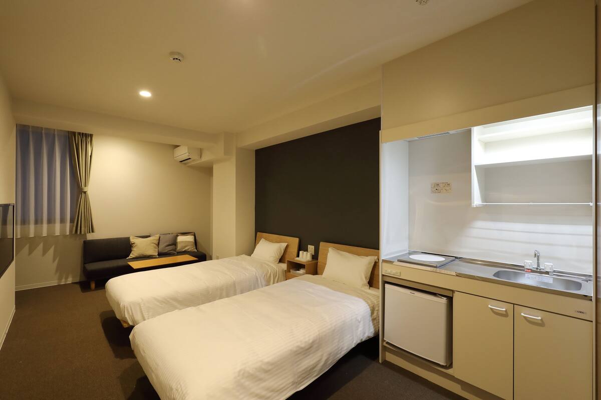 A hotel room with two beds, a small kitchenette, a sofa, and a window with curtains.