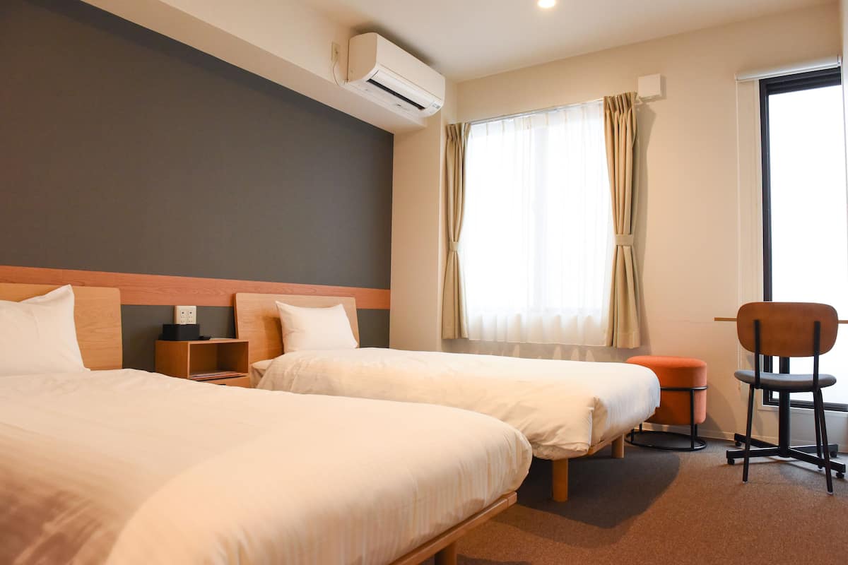 A hotel room with two beds, a chair, a small table, and a window with curtains.