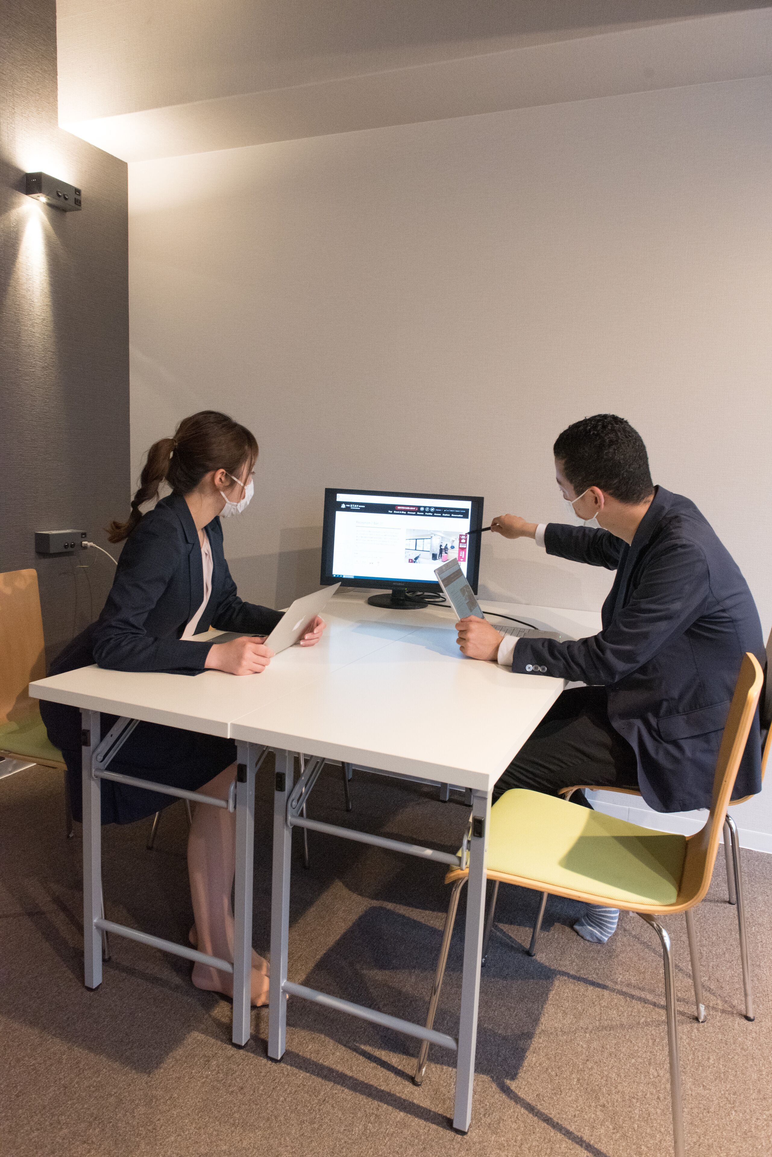 Deux personnes sont assises à une table blanche devant un écran d’ordinateur. L’une utilise une tablette, tandis que l’autre pointe du doigt l’écran.