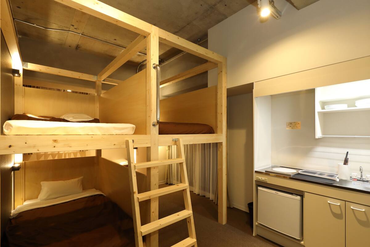 A bunk bed room with a ladder, a small kitchenette with a sink and a refrigerator, and a window with curtains.