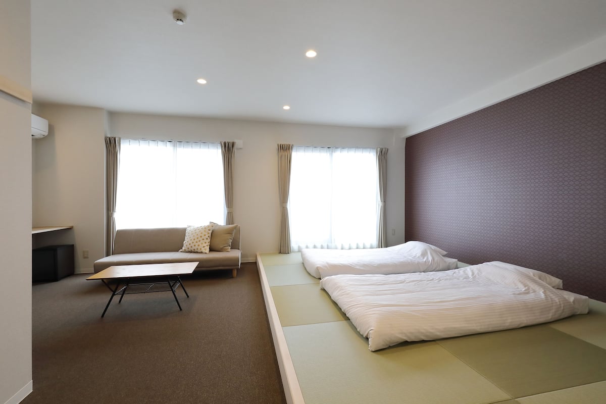 A hotel room with a sofa, two beds, a small table, and large windows.