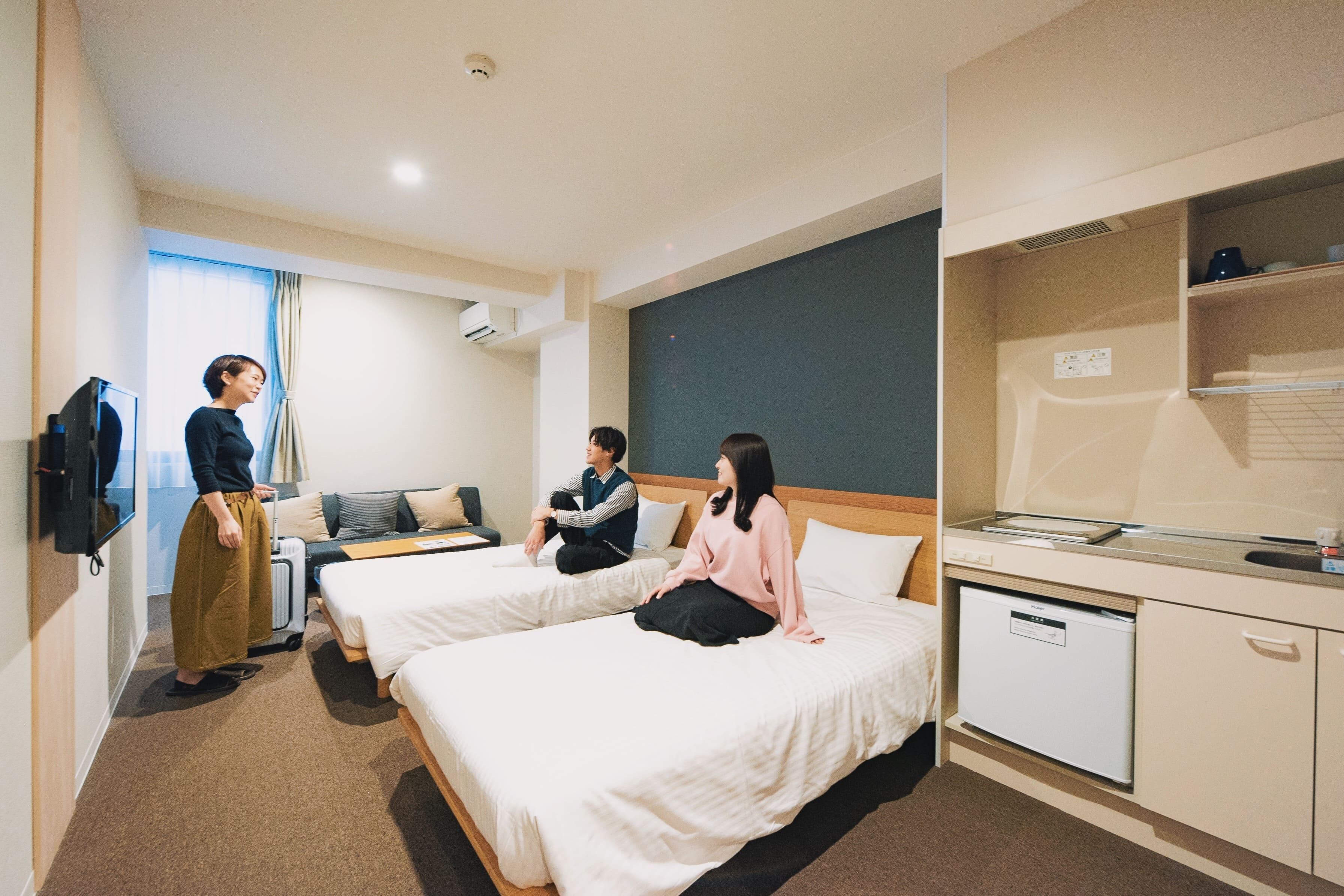 Habitación de hotel con dos camas, un escritorio, una kitchenette pequeña y una ventana con cortinas.
