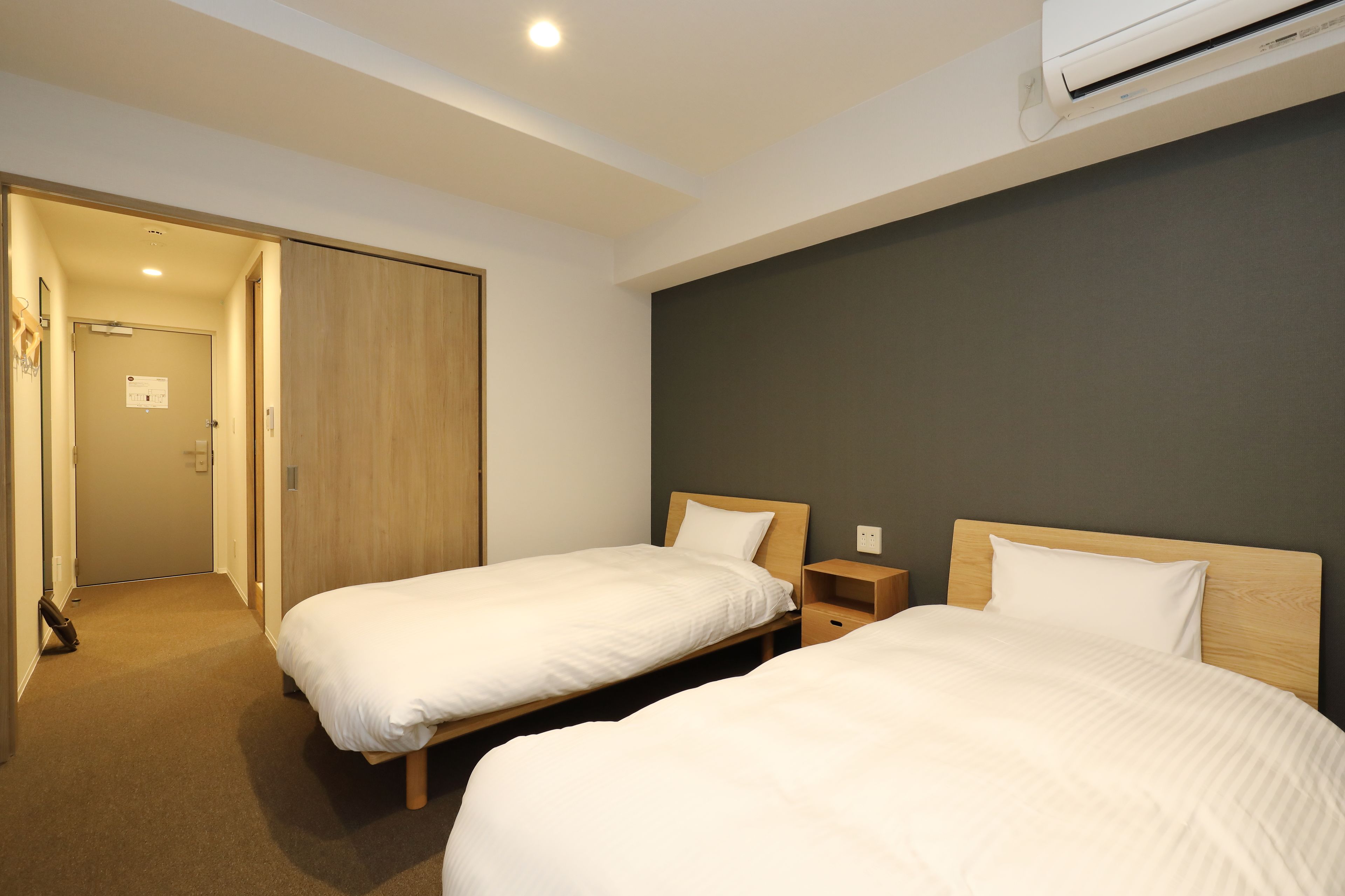 A hotel room with two single beds, a wooden wardrobe, a small bedside table, and an air conditioning unit mounted on the wall.