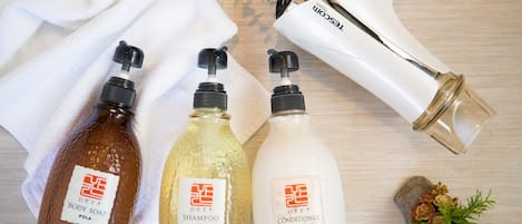 Tres botellas de productos de cuidado corporal sobre una superficie de madera, con una toalla blanca, un secador de pelo y piedras decorativas.