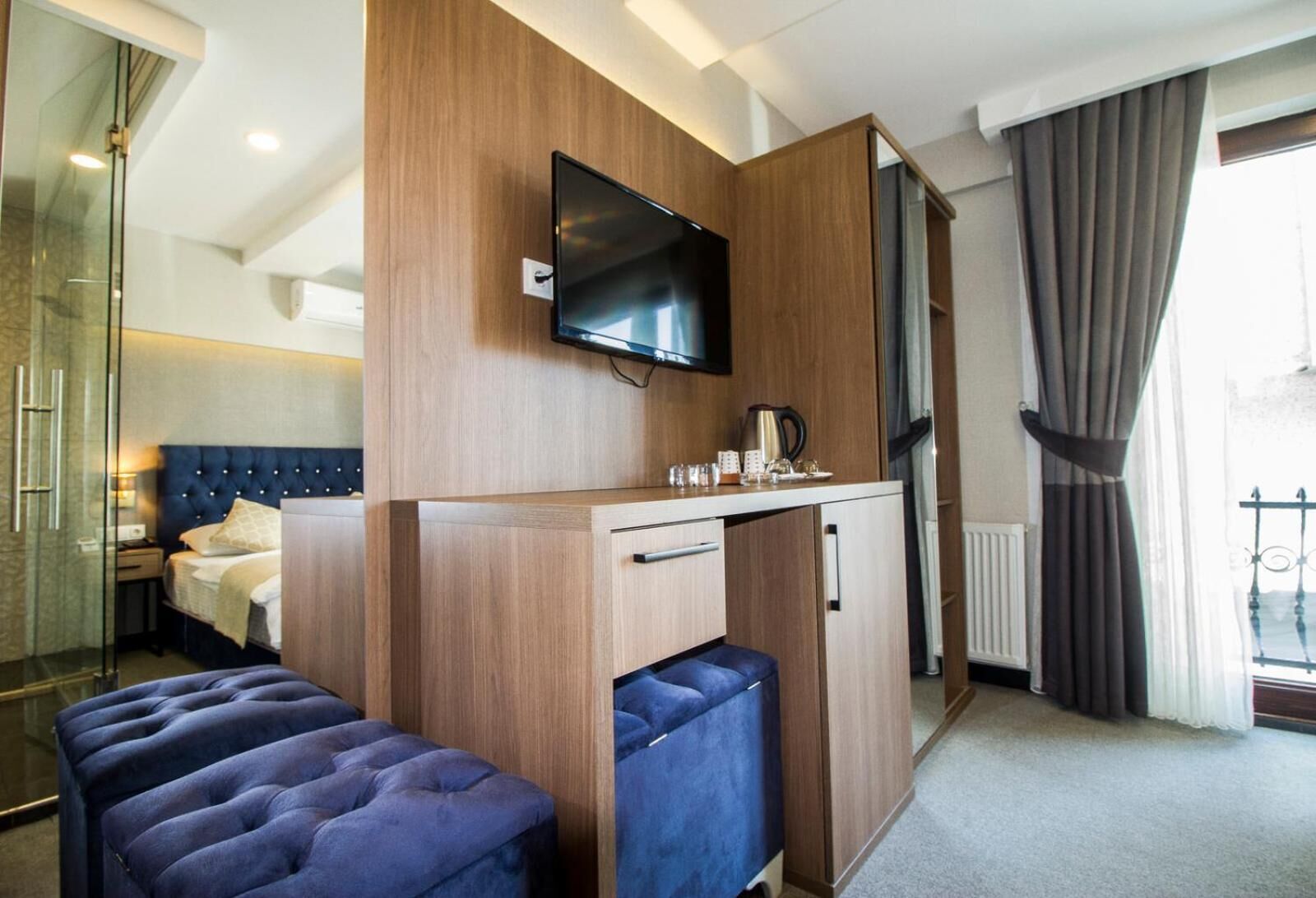 Habitación de hotel pequeña con una cama, un televisor fijado a un panel de madera, una kitchenette compacta y una zona de estar con un banco acolchado.