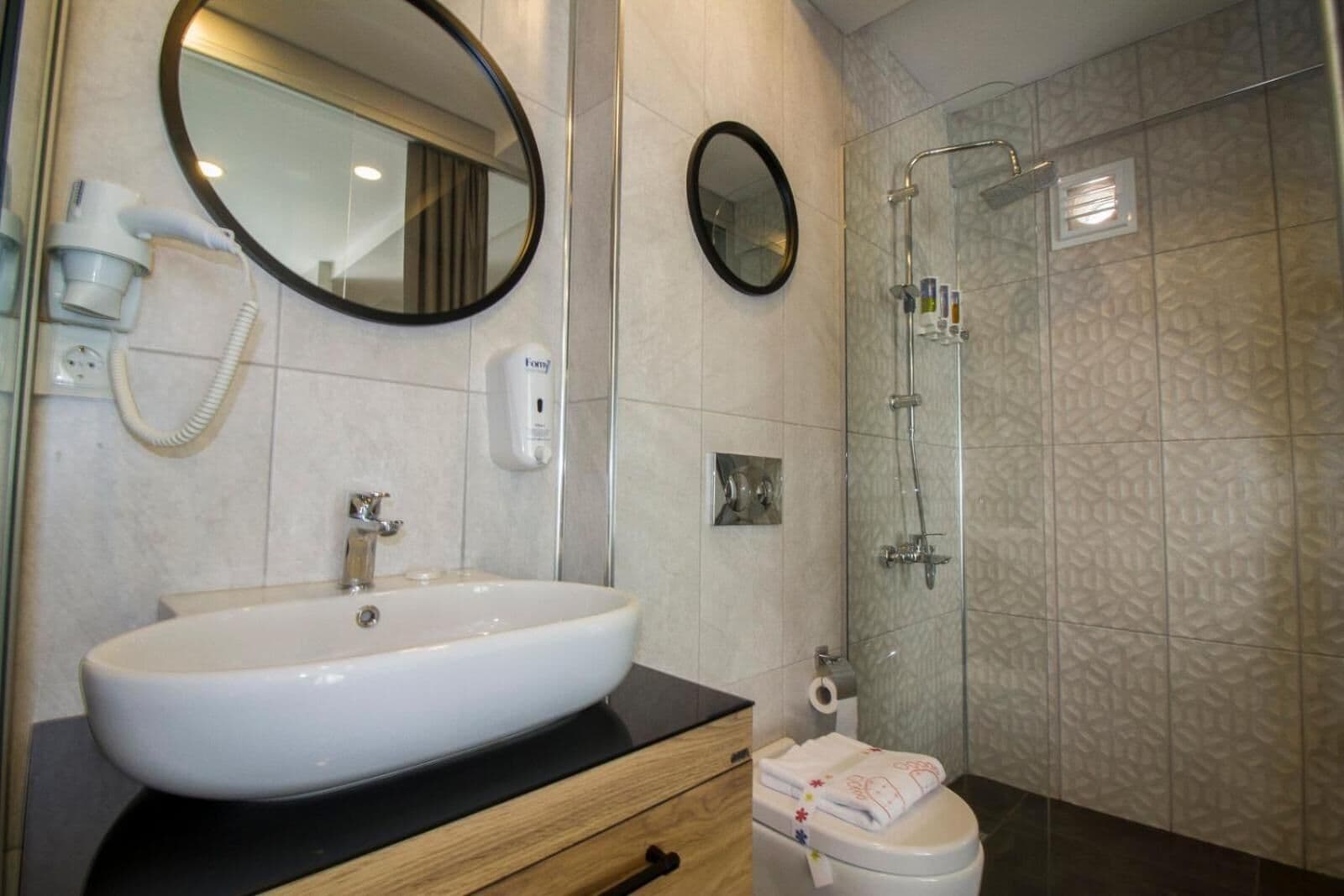 Ein Badezimmer mit einem großen, weißen Waschbecken, einem Spiegel, einem Handtrockner, einer Toilette und einer Duschkabine aus Glas.