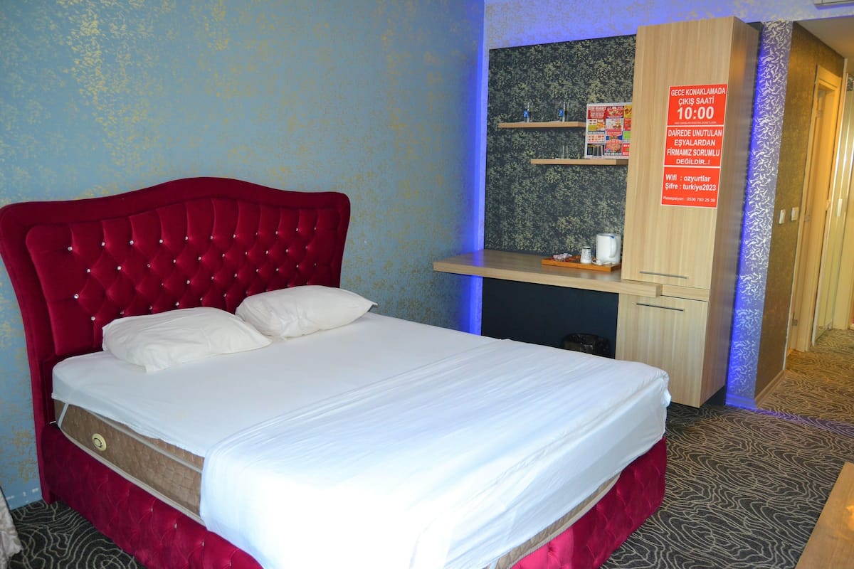 A hotel room with a red upholstered bed, a small desk with a computer, and a wall-mounted shelf with toiletries.