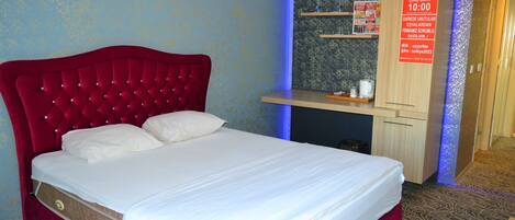 A hotel room with a red upholstered bed, a small desk with a computer, and a wall-mounted shelf with toiletries.