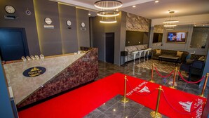 Un espace d’accueil d’hôtel avec un comptoir en marbre, des horloges, un téléviseur et un tapis rouge menant à un coin salon.