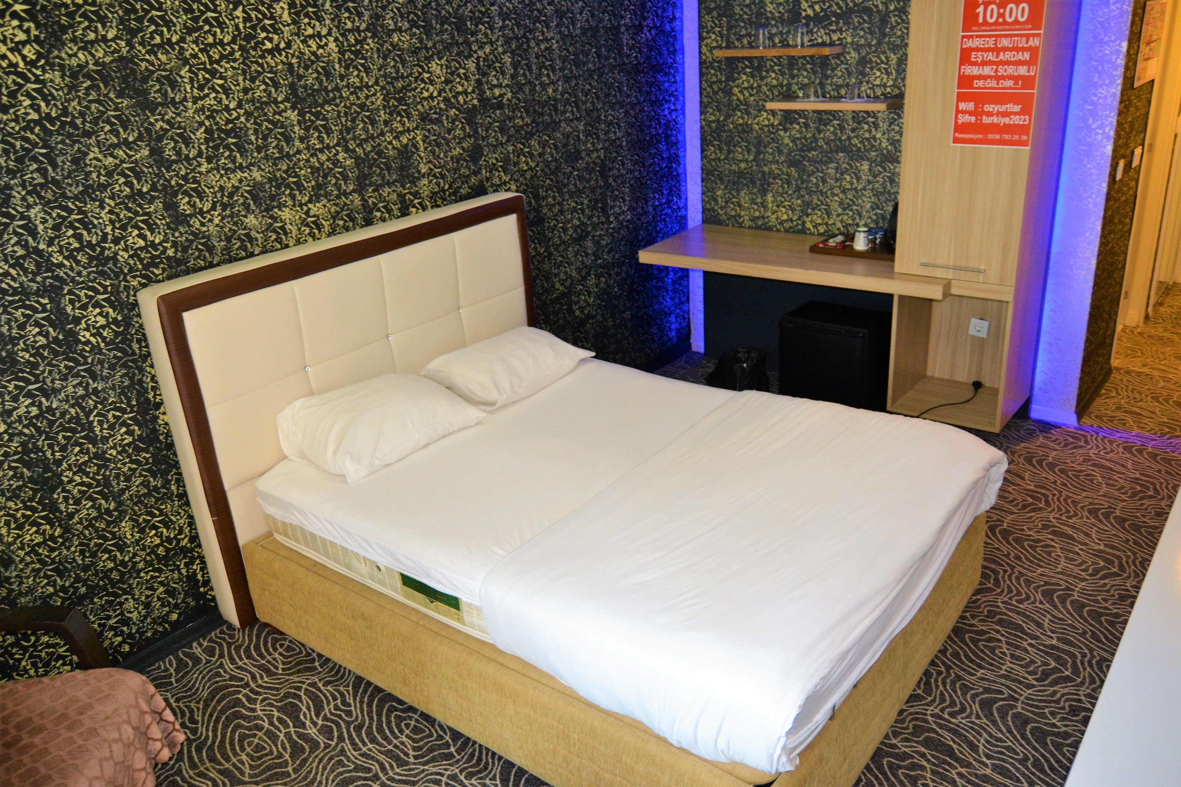 A neatly made bed with a wooden headboard in a room with patterned wallpaper and a small desk with a shelf.