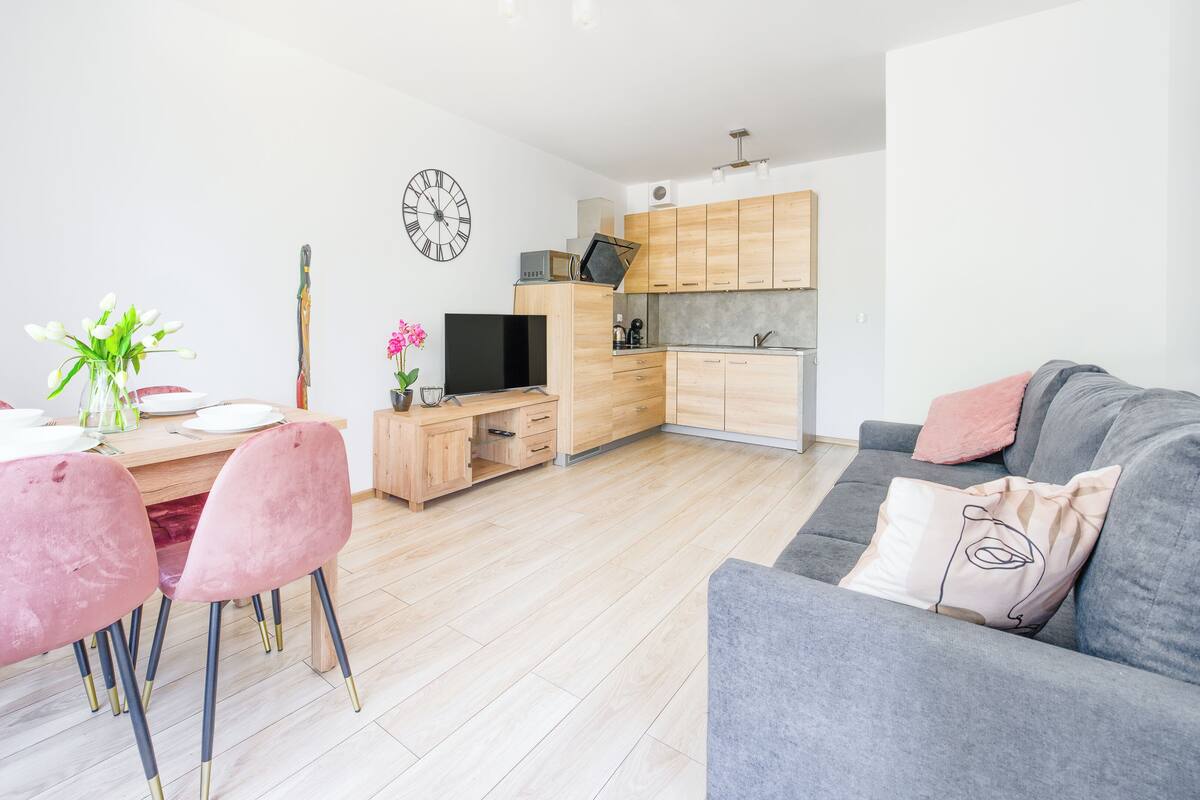 Ein modernes Wohnzimmer mit grauem Sofa, rosafarbenen Stühlen, einem Holzespeisetisch mit Tellern, einem Fernseher und einer Küchenzeile mit Holzschränken und einer Uhr an der Wand.