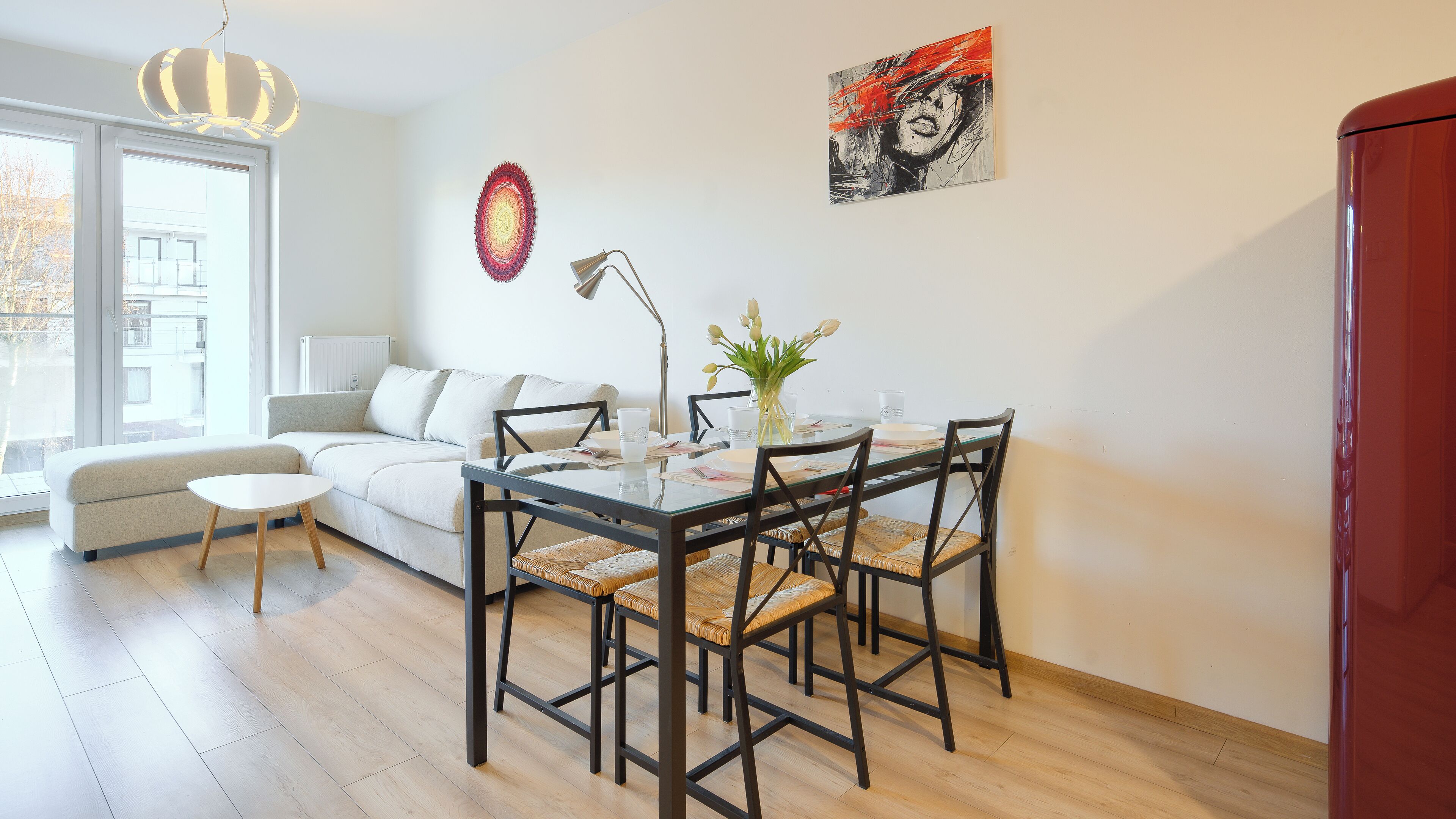 Una sala moderna con una mesa de vidrio, un sofá y una mesa de centro. Hay un cuadro en la pared y una ventana con vistas a edificios.
