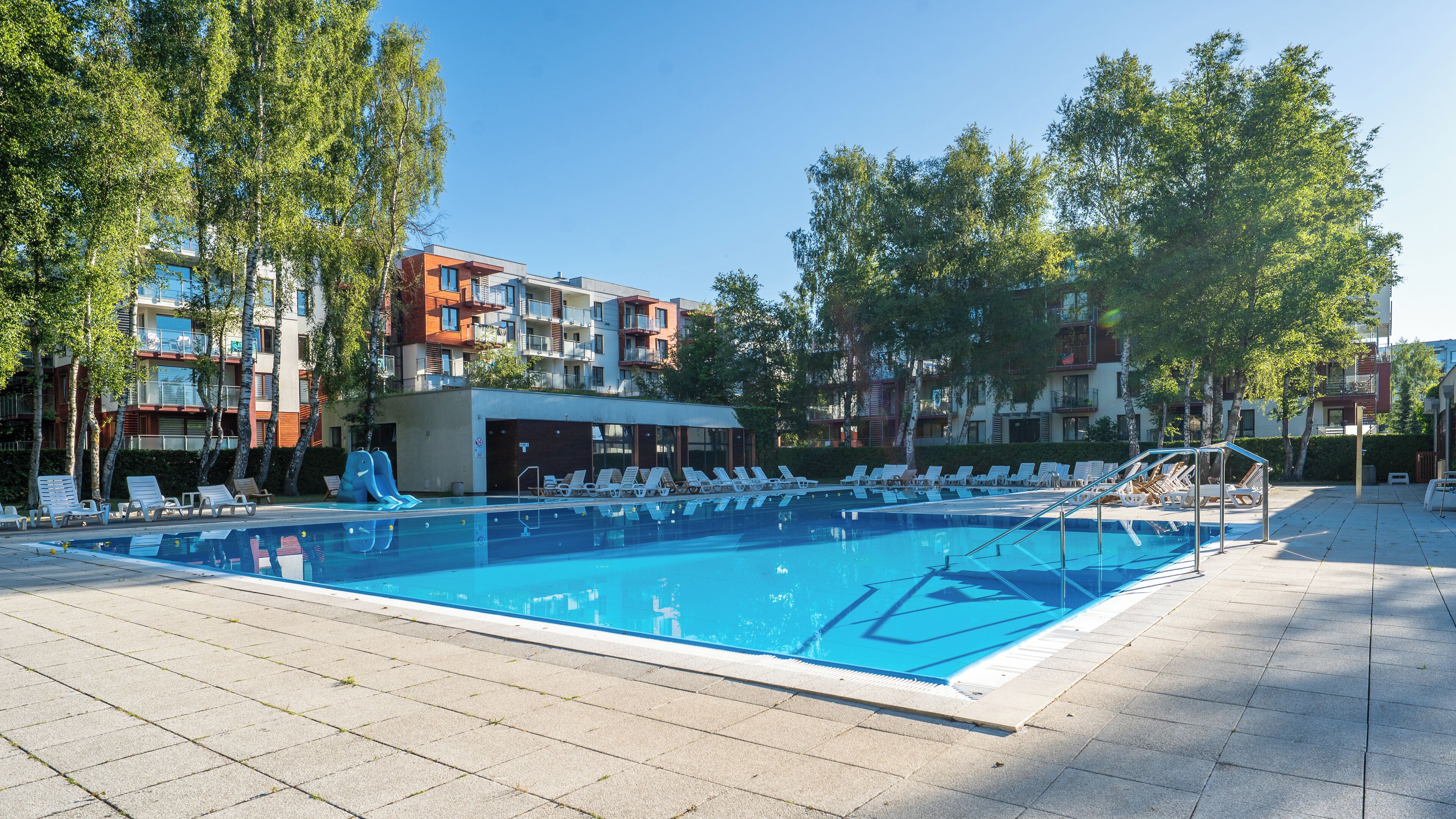 Une piscine entourée d’arbres et d’immeubles à plusieurs étages, avec des chaises longues à proximité.