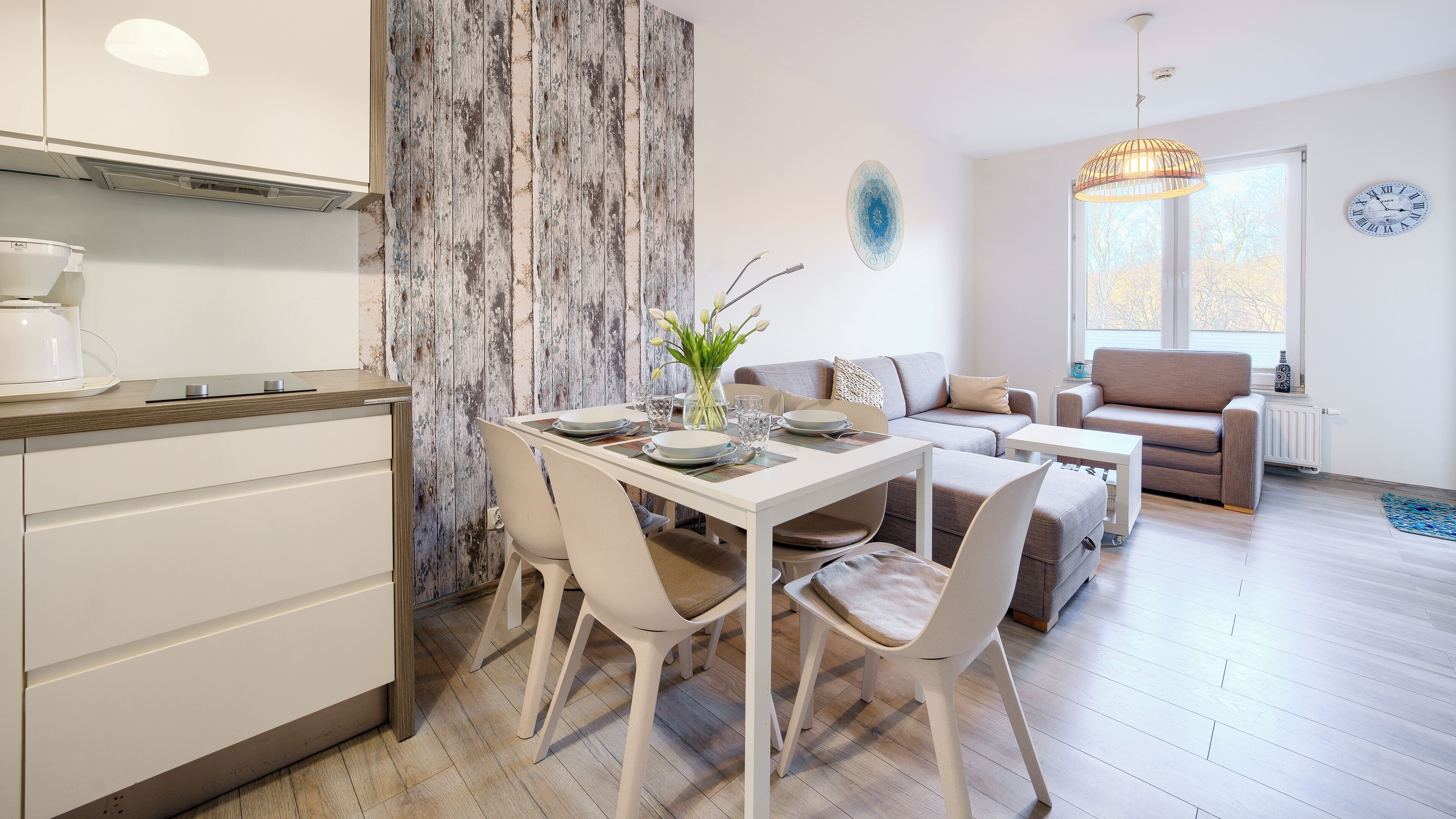 A modern kitchen with white cabinets, a dining table set with plates and glasses, and a living area with a sofa and a window with a view.