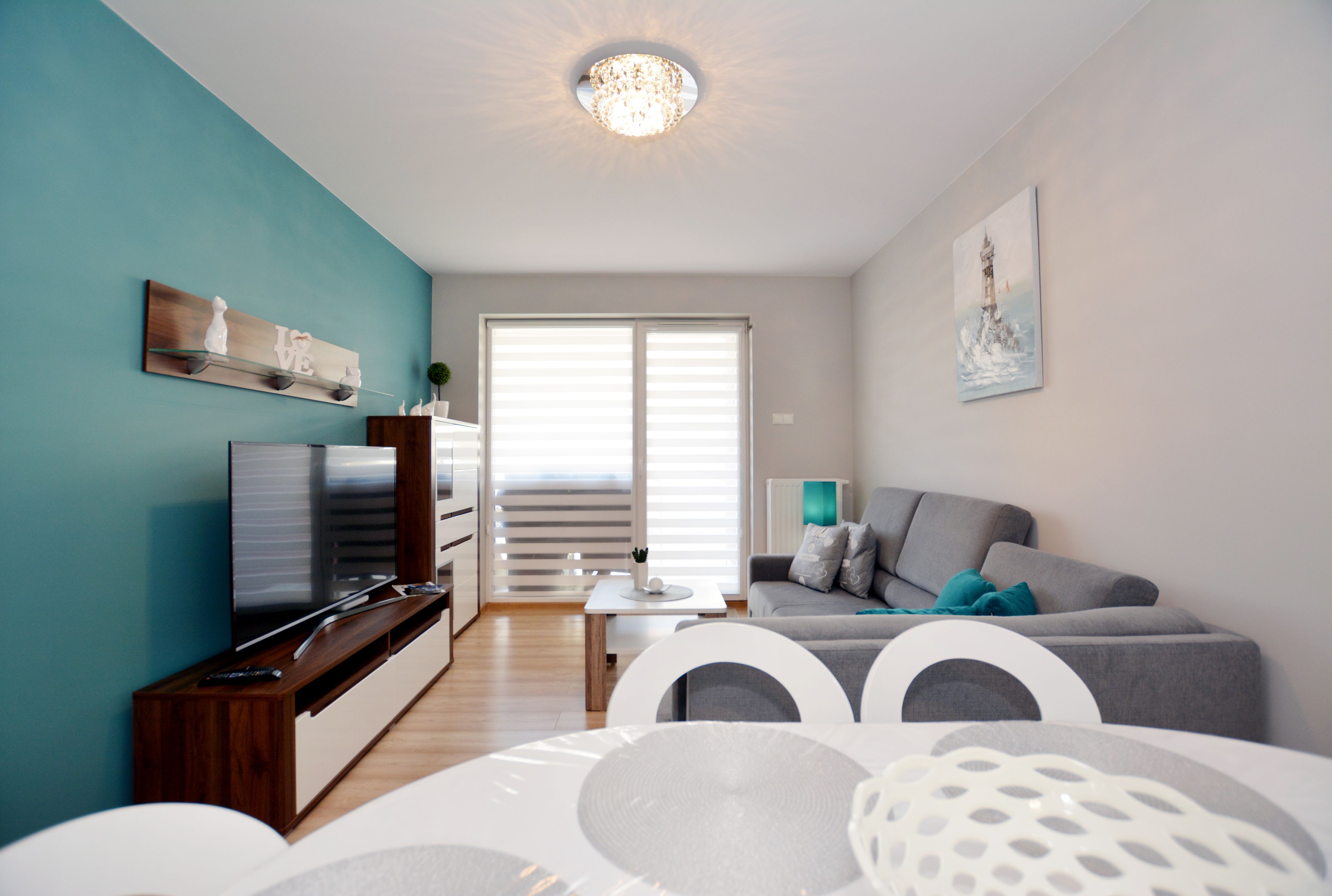 A modern living room with a flat-screen TV, a grey sofa, a white dining table, and teal walls.
