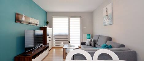 A modern living room with a flat-screen TV, a grey sofa, a white dining table, and teal walls.