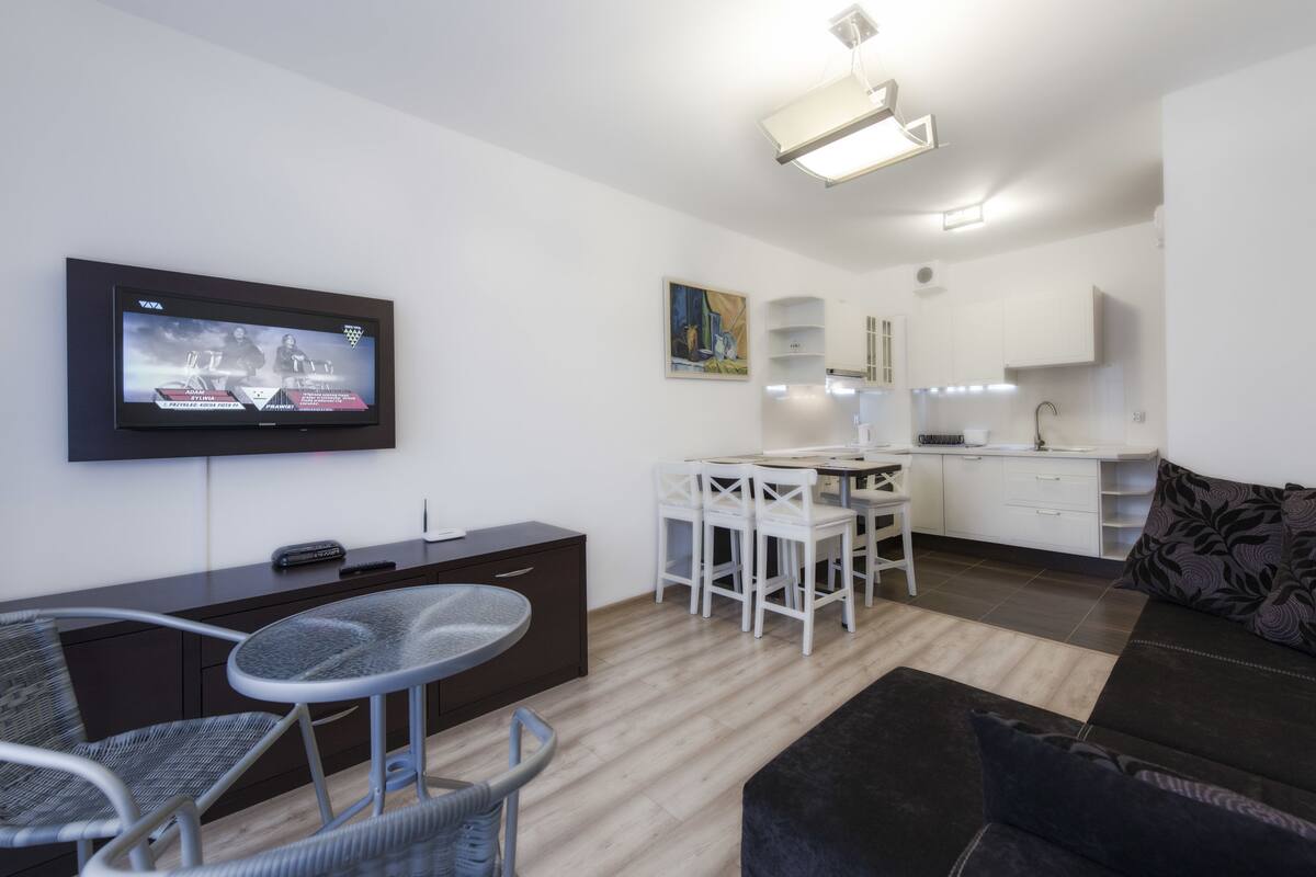 Ein modernes Wohnzimmer mit Flachbildfernseher, einem Essbereich mit weißen Stühlen, einer Küche mit weißen Schränken und einem schwarzen Sofa.