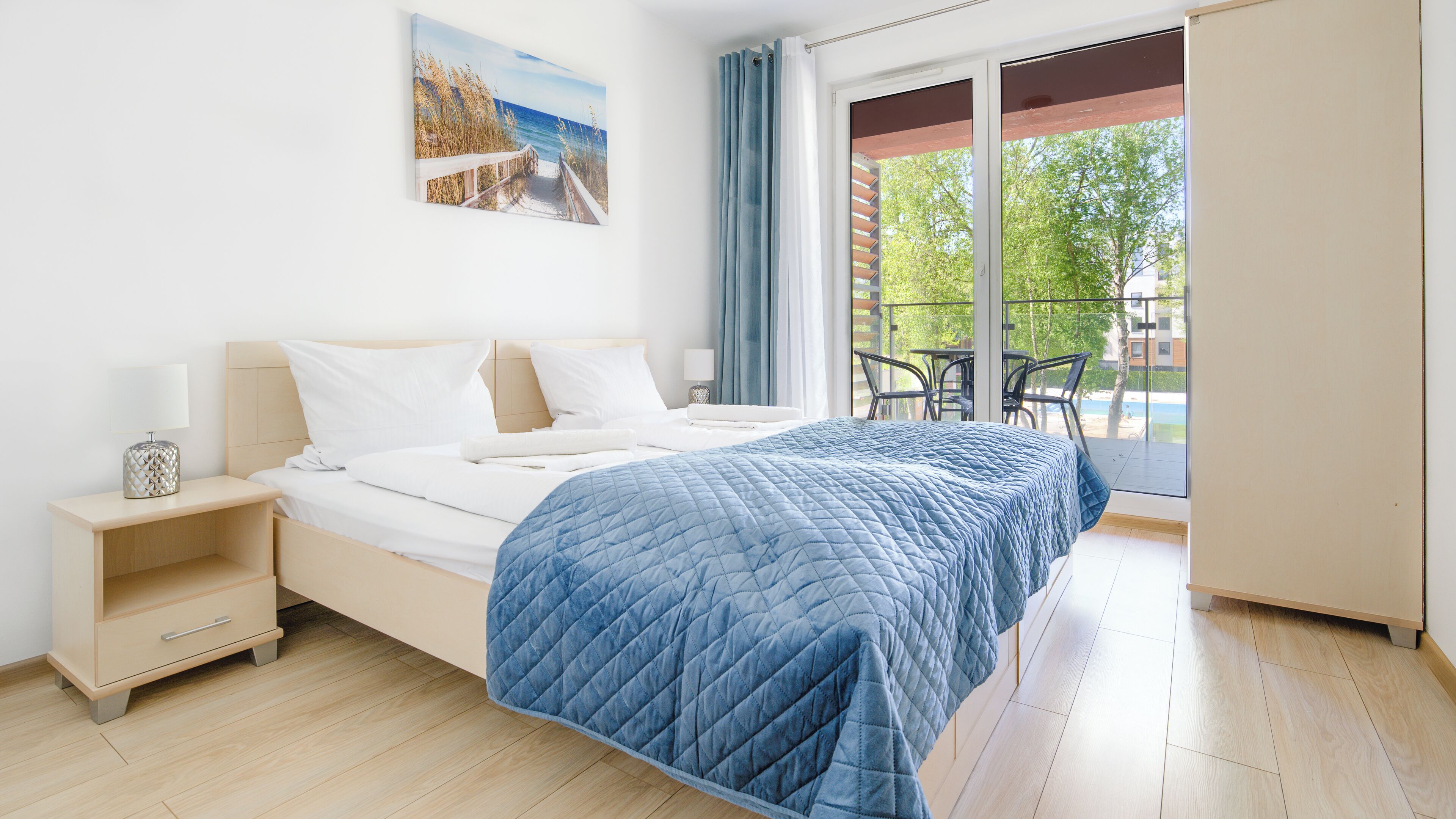 Una habitación de hotel moderna con una cama grande, una mesita de noche, una lámpara y vistas al exterior a través de un ventanal corredero.
