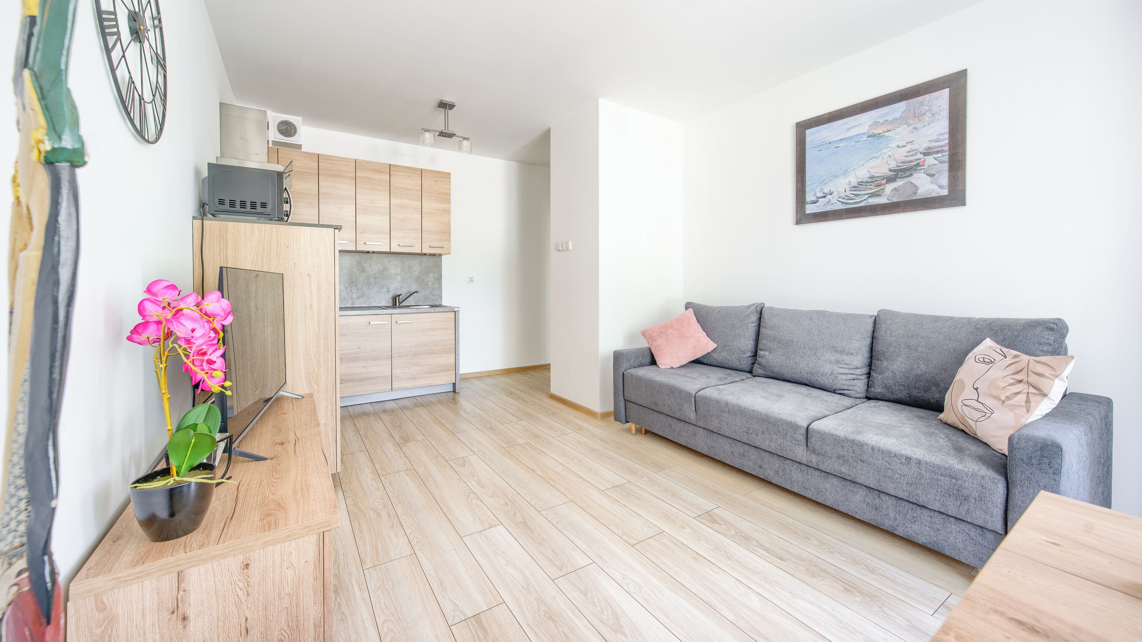 A modern living room with a grey sofa, a small kitchen area, and a wooden floor.
