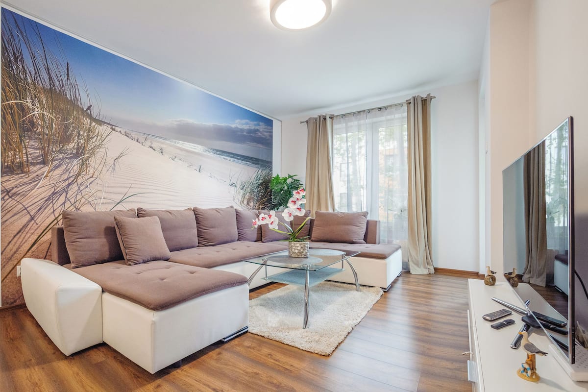 Ein modernes Wohnzimmer mit einer Eckcouch, einem Glastisch und einem großen Wandbild mit Strandmotiv.