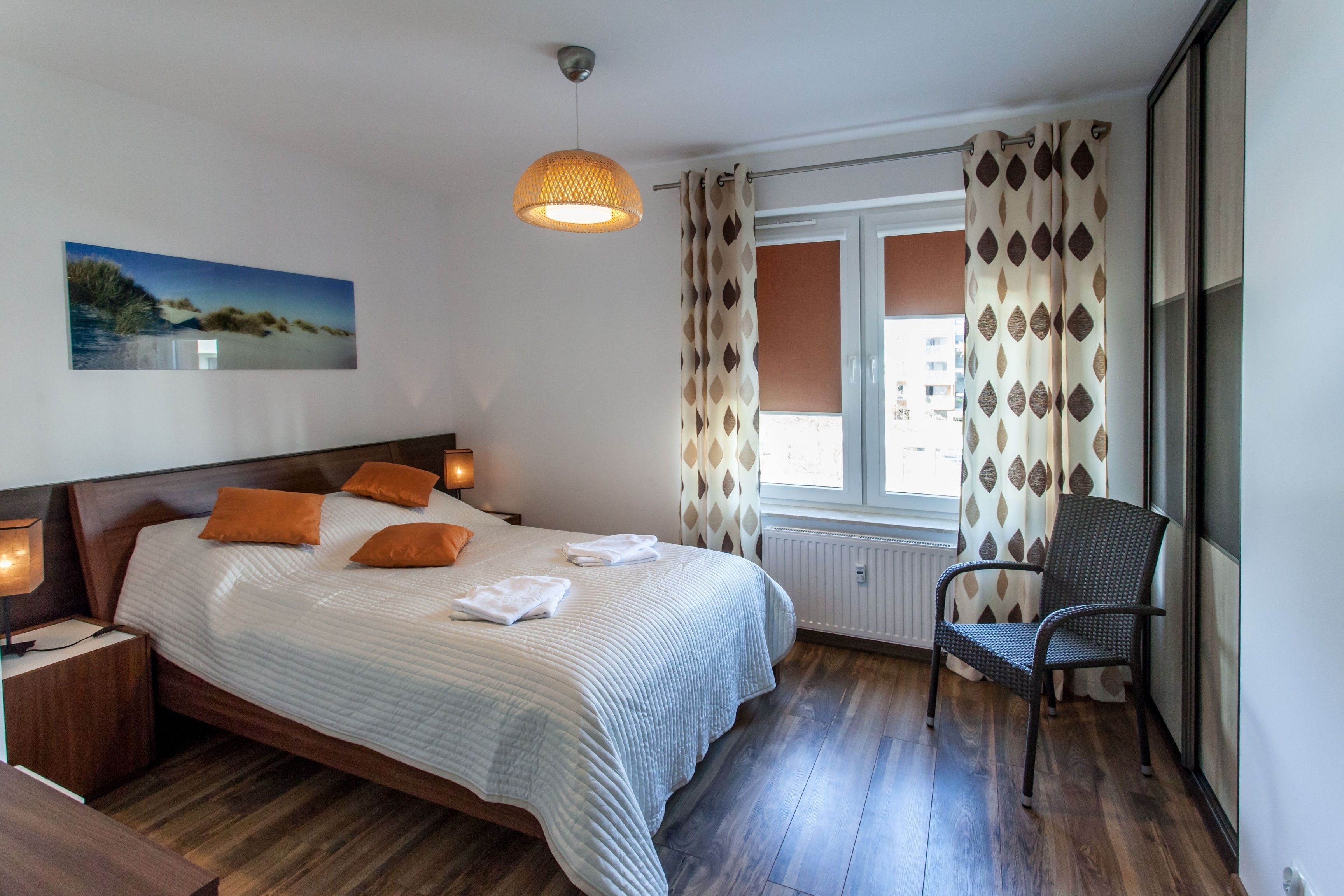 A bedroom with a bed, bedside lamps, a chair, a window with curtains, and a wall-mounted picture.