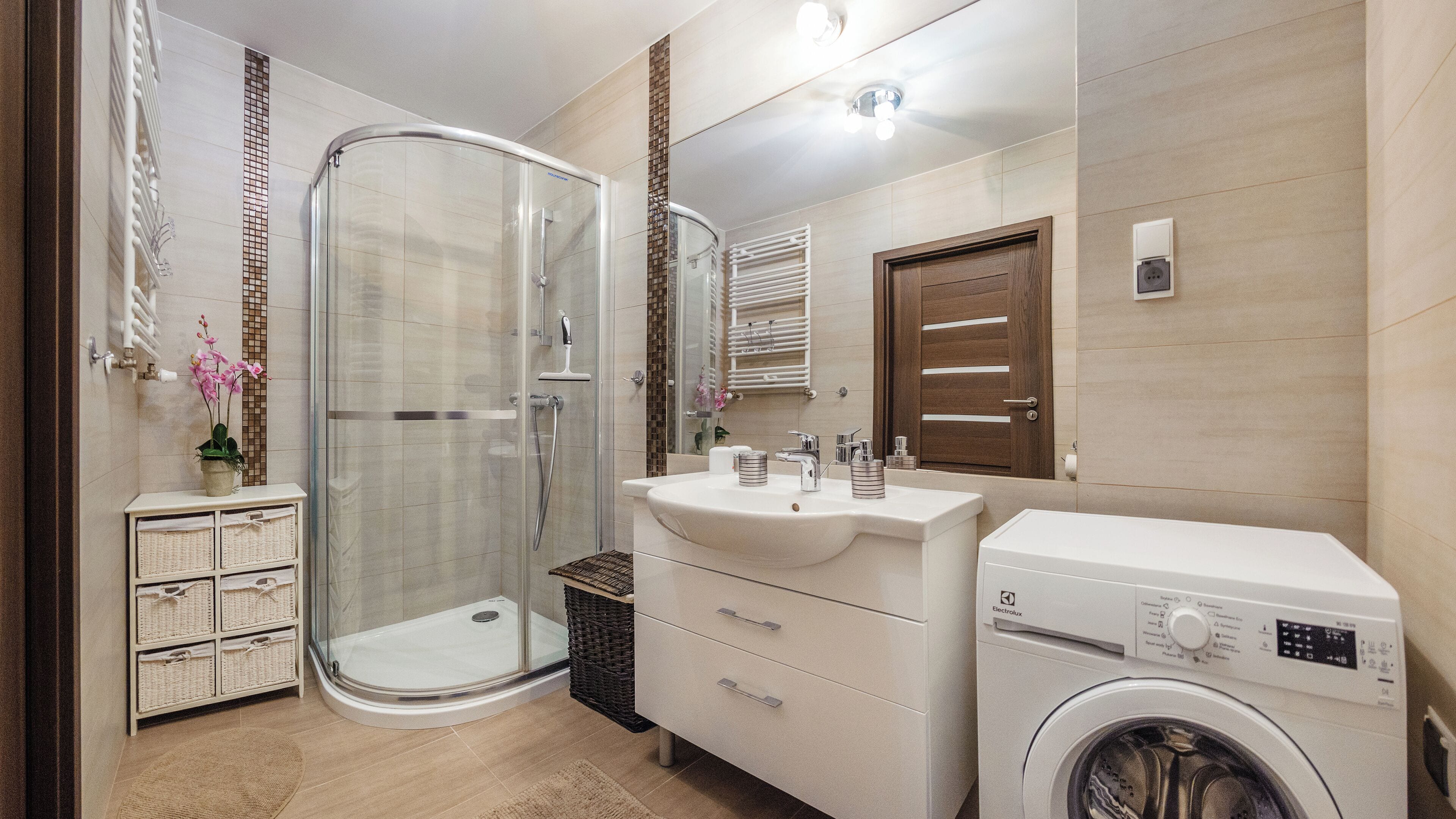 Un baño moderno con ducha de vidrio, un lavamanos blanco con espejo y una lavadora de carga frontal.