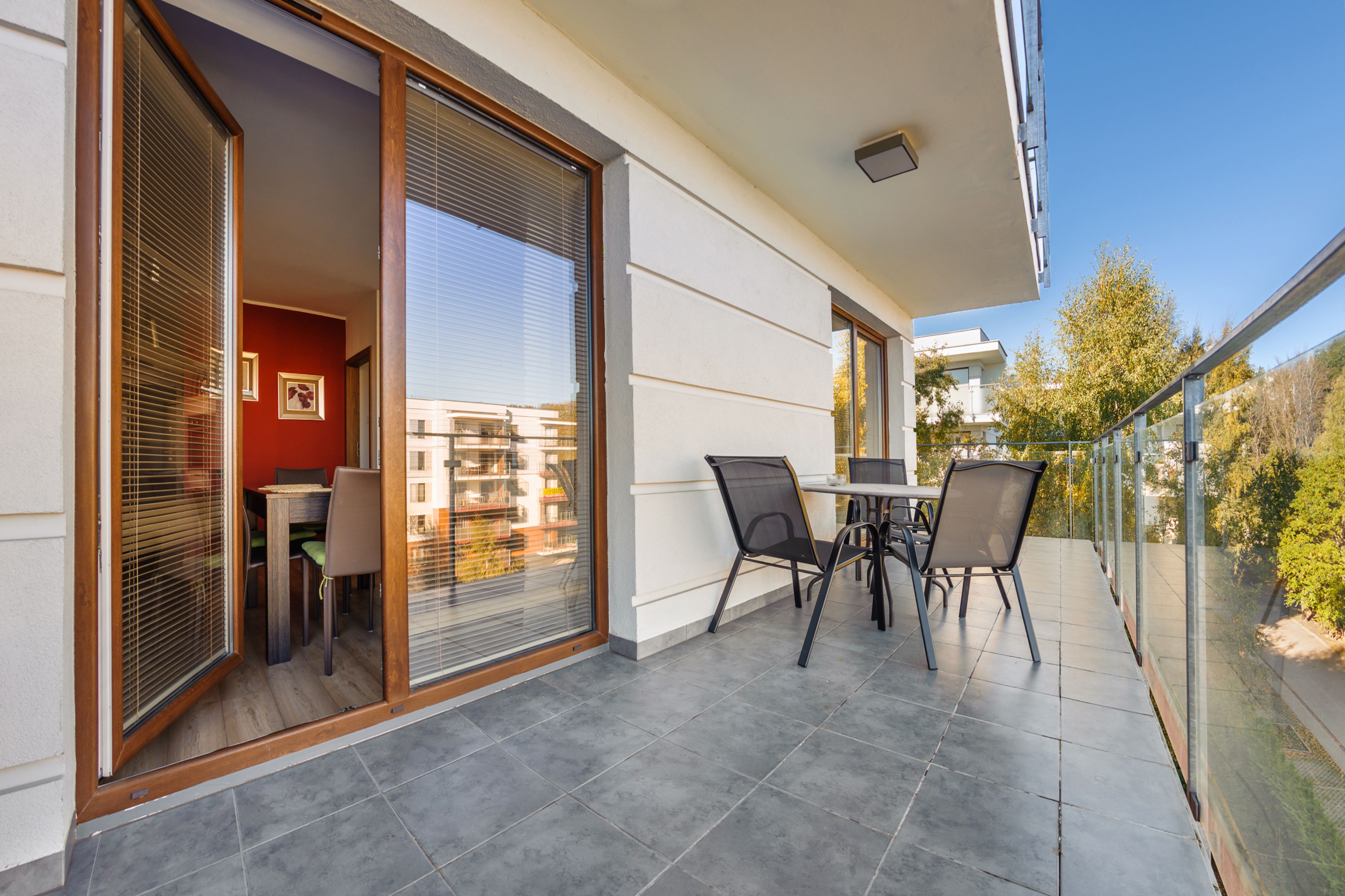 A balcony with a table and chairs, a sliding glass door, and a view of a building and trees.
