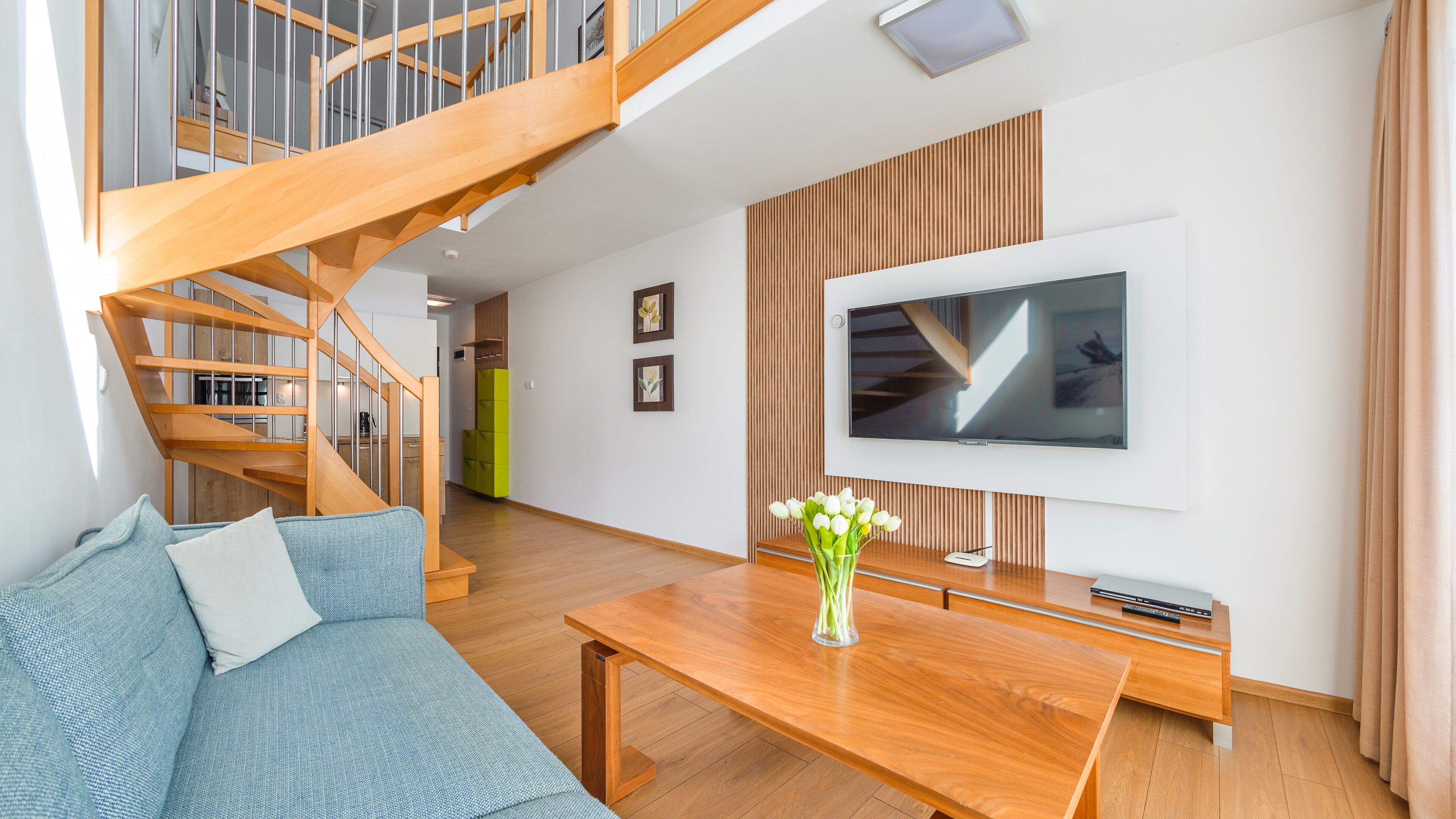 A modern living room with a wooden staircase, a flat-screen TV, a wooden coffee table, and a blue sofa.