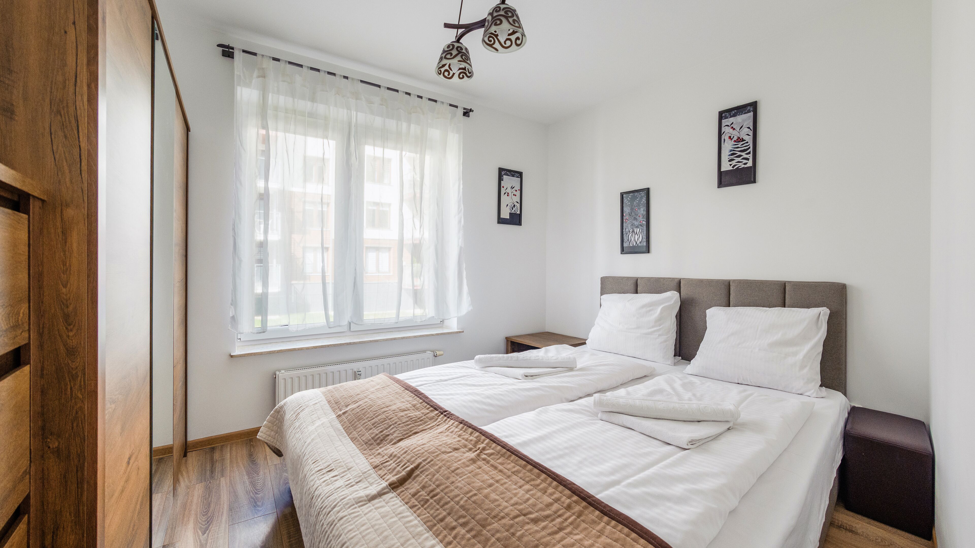 Une chambre à coucher avec un grand lit, une table de chevet, une commode, un radiateur et une fenêtre avec des rideaux légers.