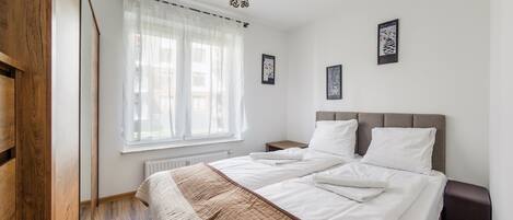 A bedroom with a large bed, a bedside table, a nightstand, a radiator, and a window with sheer curtains.