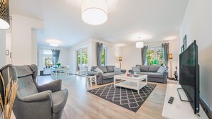 A modern living room with a grey sofa, a white coffee table, and a flat-screen TV. There is a dining area with a table and chairs, and a large window with curtains. - Apartamenty Sun & Snow Polanki (Kolobrzeg)