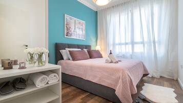 A bedroom with a bed, a shelf with towels, a vase with flowers, and a painting on the wall.