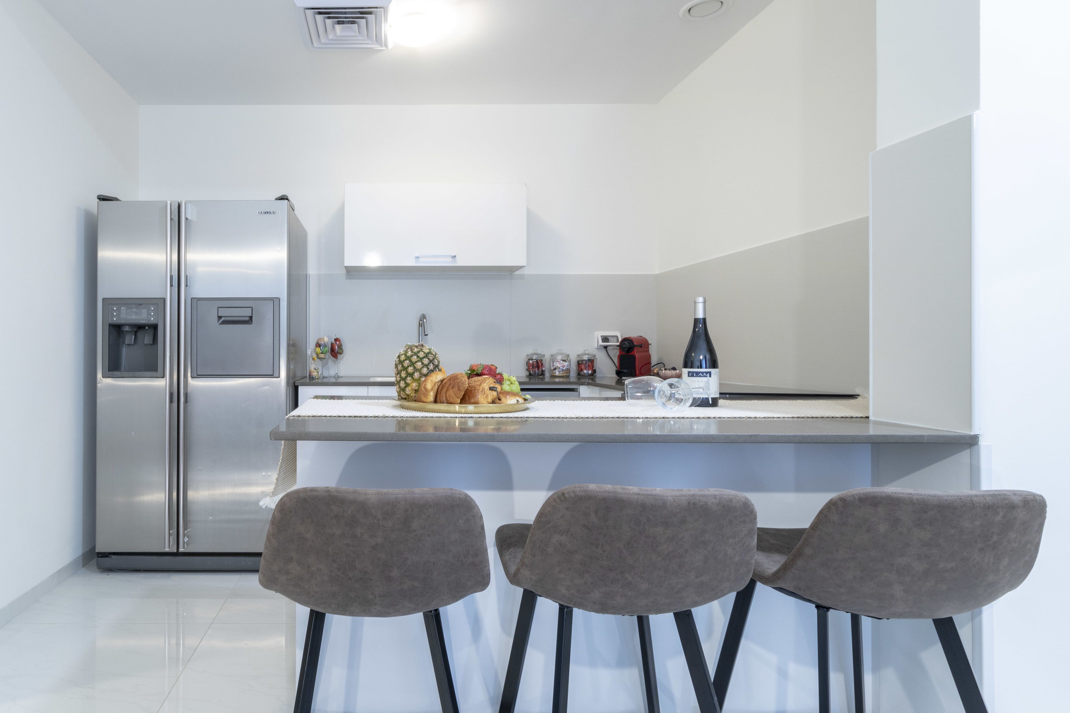 A modern kitchen with a stainless steel refrigerator, a white cabinet, a small island with a fruit bowl and a bottle, and two bar stools.
