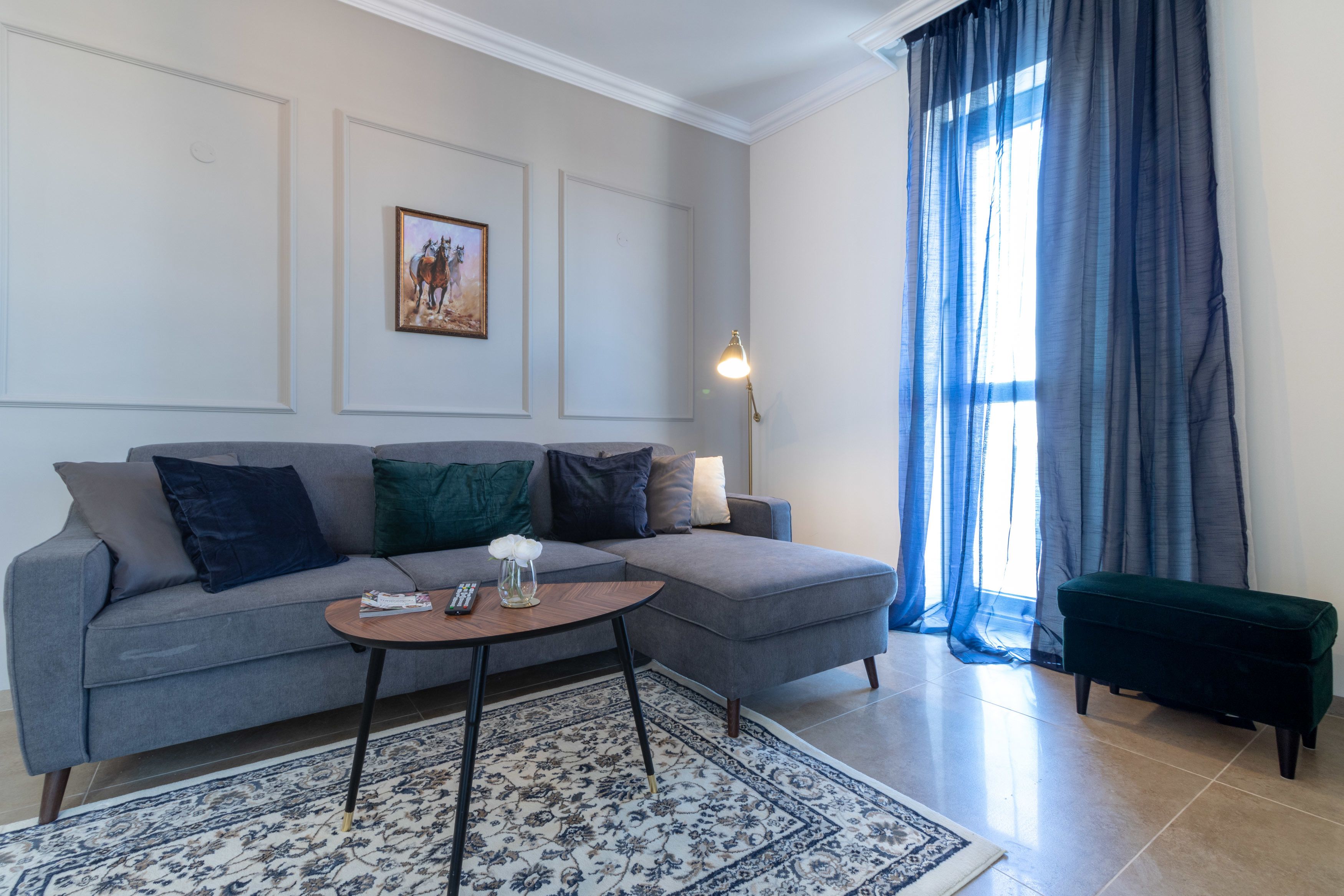 Ein modernes Wohnzimmer mit einem grauen Ecksofa, einem runden Couchtisch und einem dunkelgrünen Hocker. An der Wand hängt ein gerahmtes Bild und in der Nähe des Fensters steht eine Stehlampe.