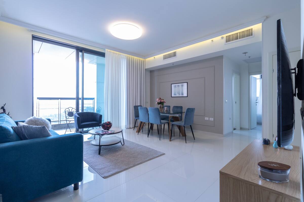 Uma sala de estar moderna com um sofá azul, uma área de jantar com cadeiras azuis e uma janela grande com cortinas.