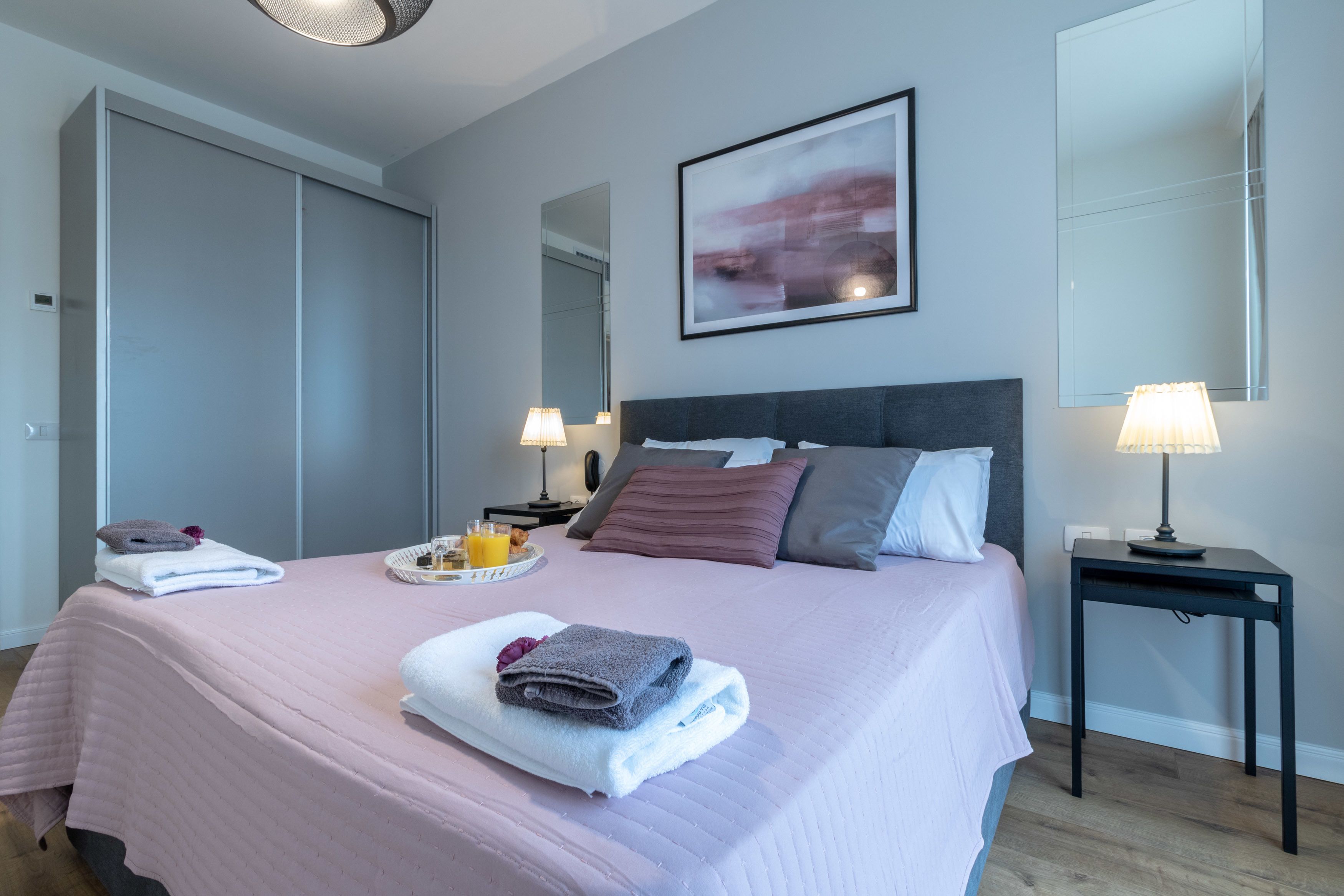 A neatly made bed with pillows, a bedside table with a lamp, a tray with breakfast items, and a large mirror on the wall.