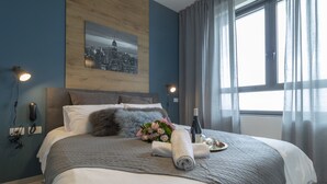 A neatly made bed with a gray blanket, a rolled towel, a bouquet of flowers, and a bottle of wine on a tray. - JTower - Isrentals (Jerusalem)