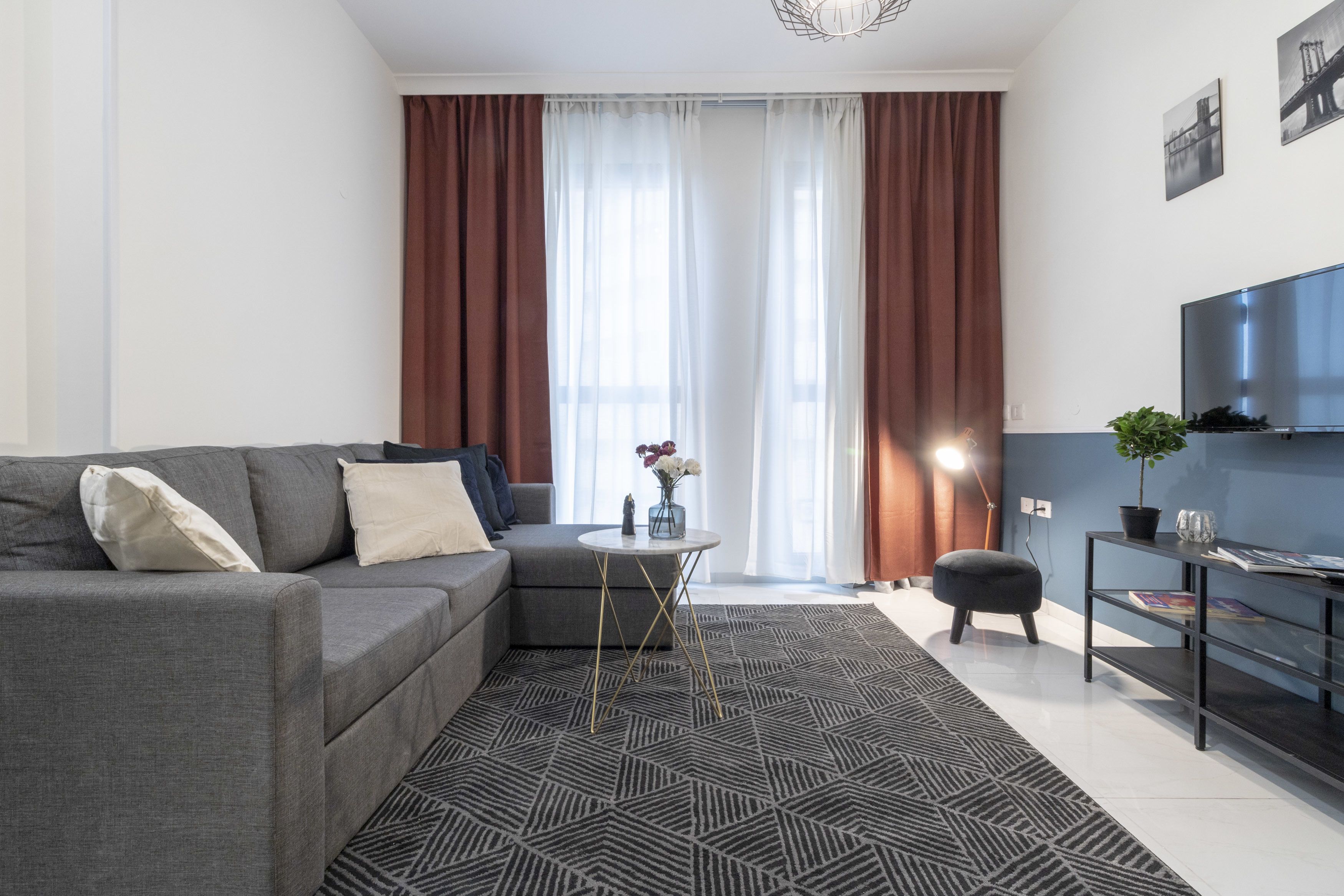 A modern living room with a grey sofa, a small round table with a vase, a black ottoman, a TV on the wall, and a large window with curtains.