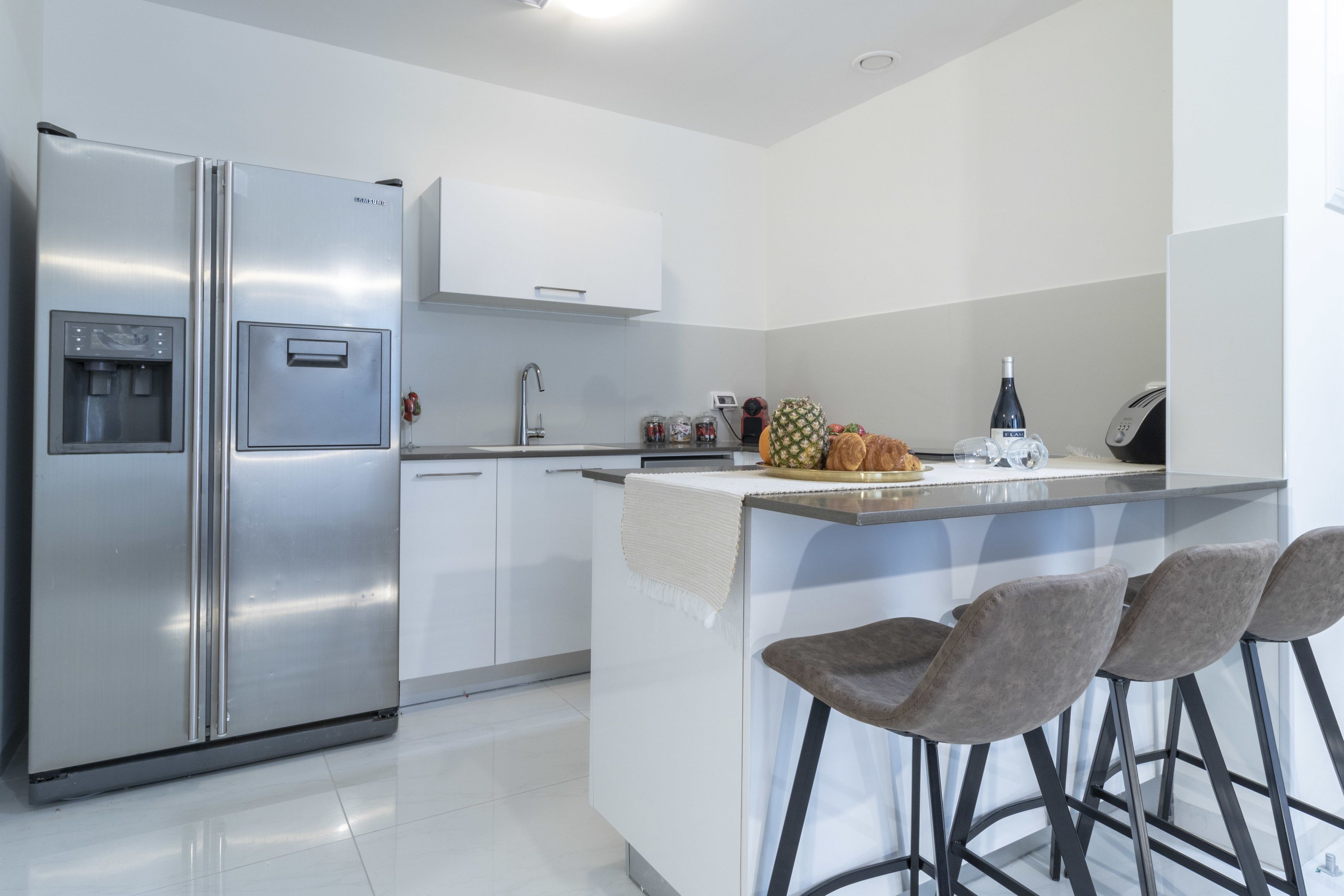 A modern kitchen with a stainless steel refrigerator, a built-in water dispenser, a sink, and a bar area with stools.