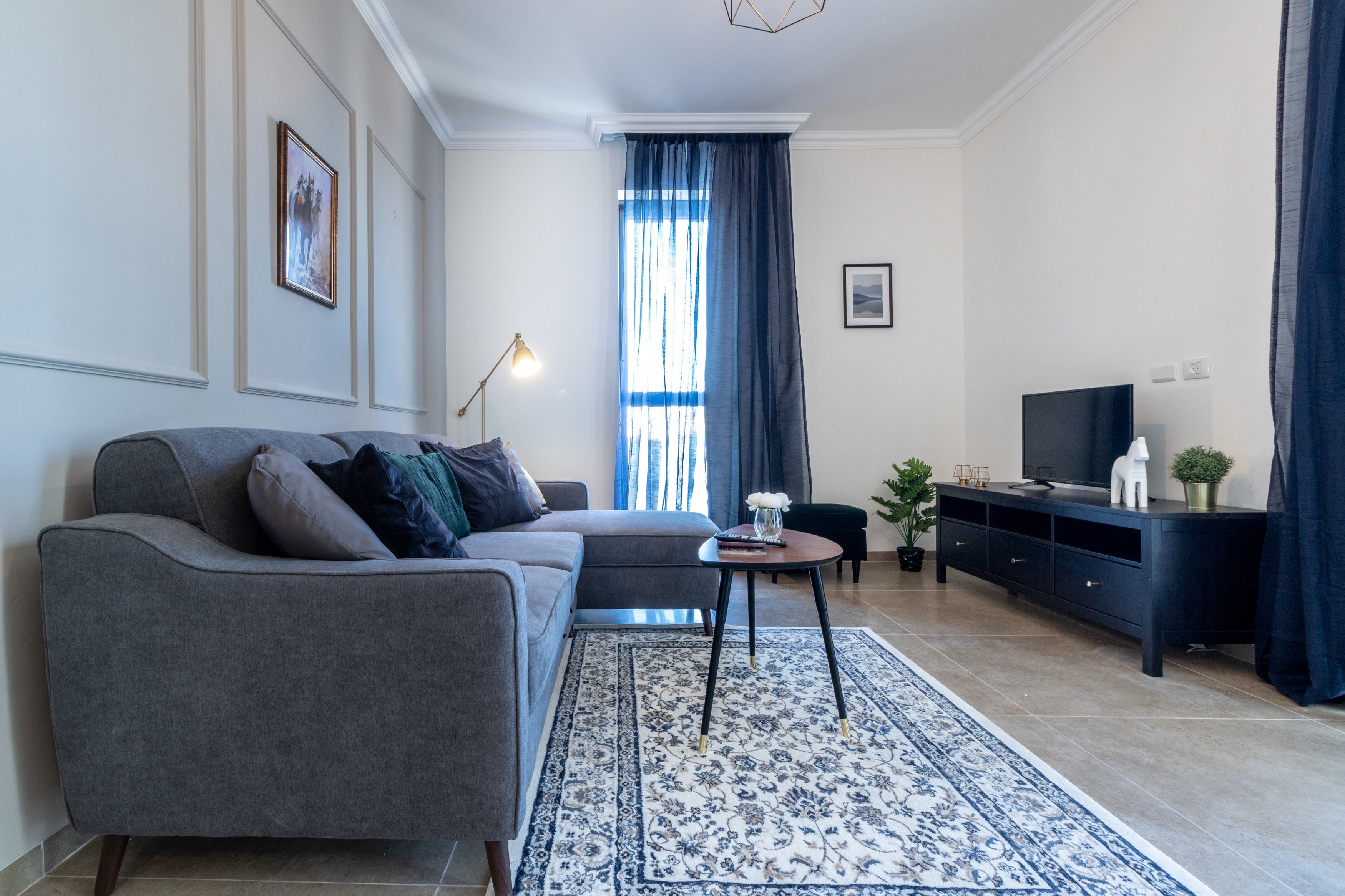 A modern living room with a grey sofa, a small round table, a TV on a black stand, and a blue patterned rug.