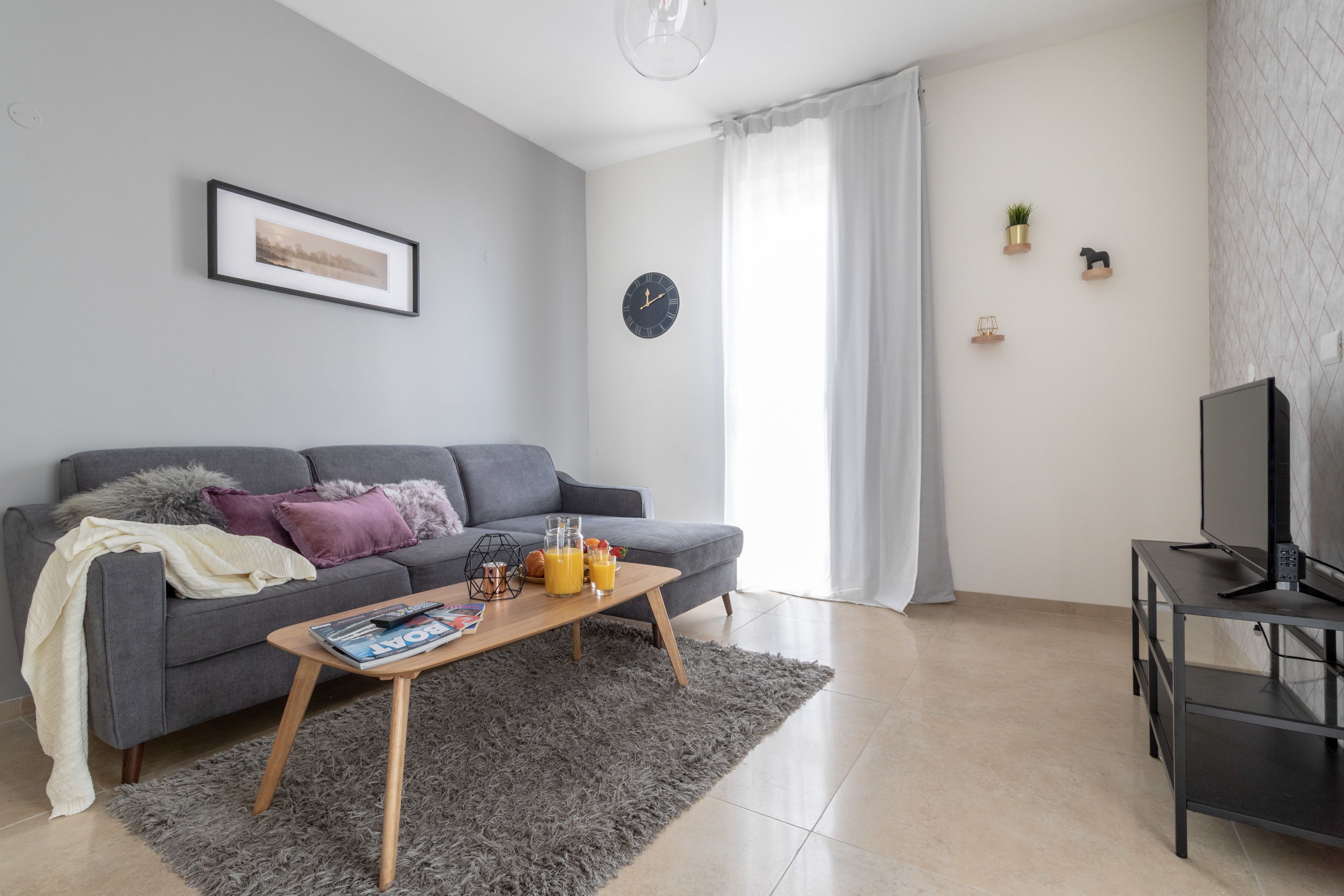A modern living room with a grey sofa, a coffee table with magazines, a TV on a stand, and a clock on the wall.
