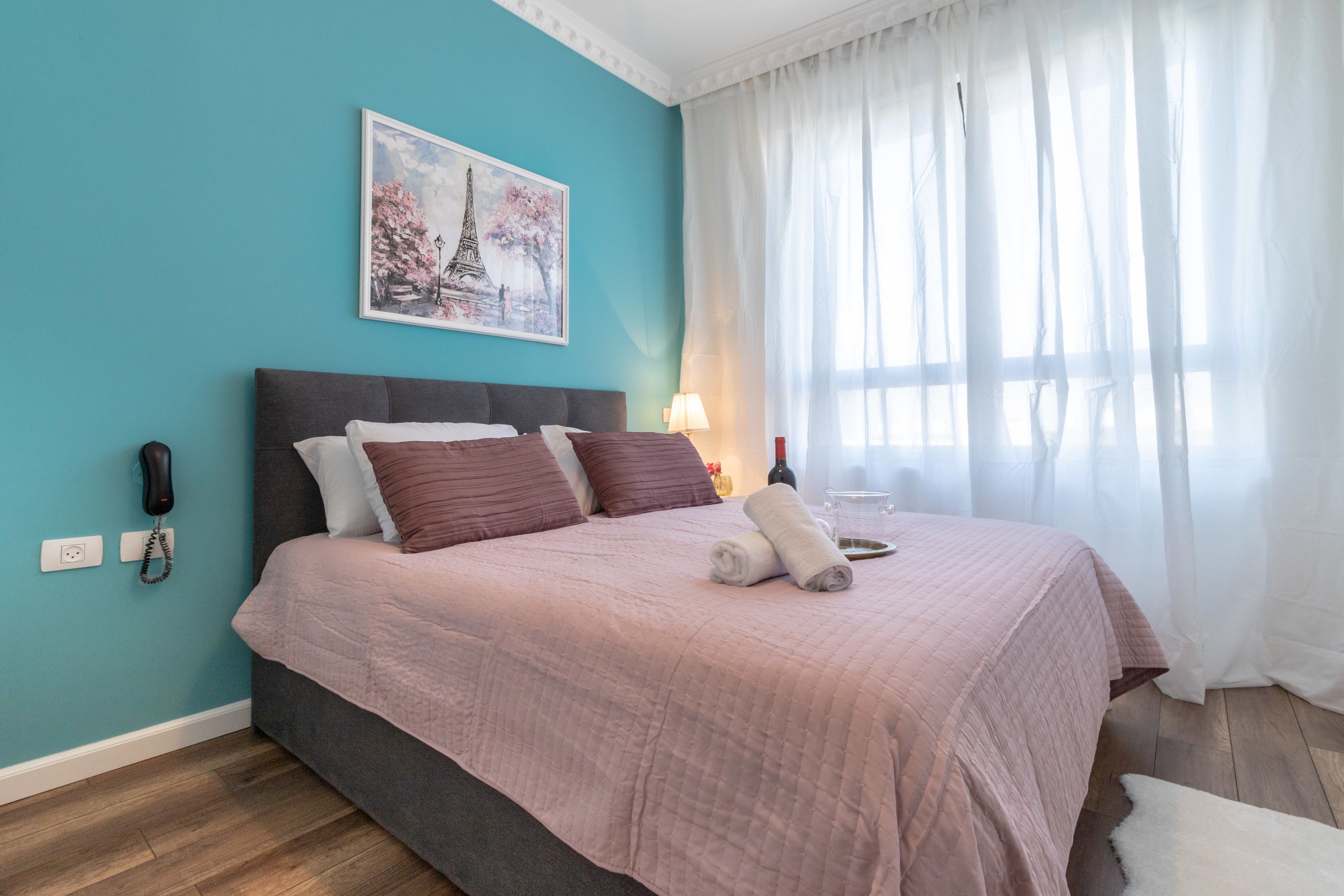 A bedroom with a bed, a telephone, a lamp, and a painting of the Eiffel Tower.